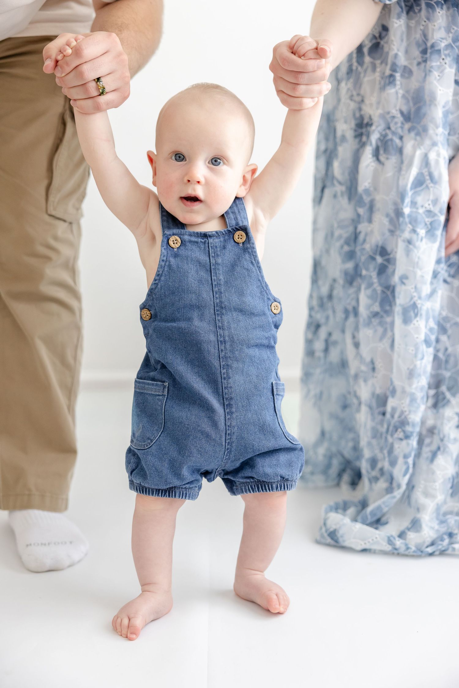 Adorable baby in denim overalls takes first steps while parents hold hands in bright white studio.