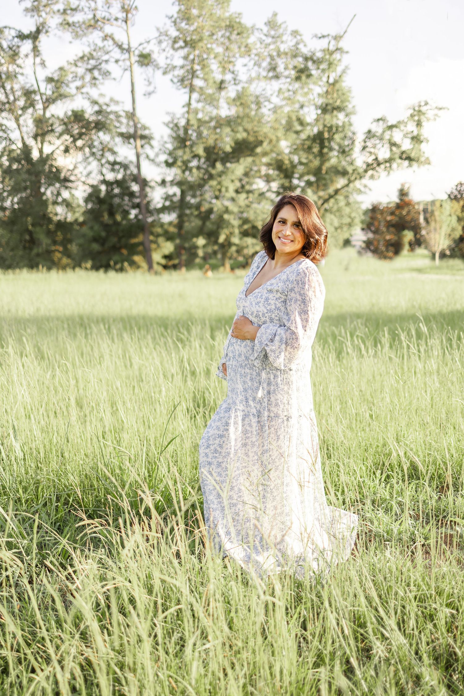 A pregnant woman in a flowing white floral dress poses gracefully in a sunlit meadow during golden hour at her maternity session.