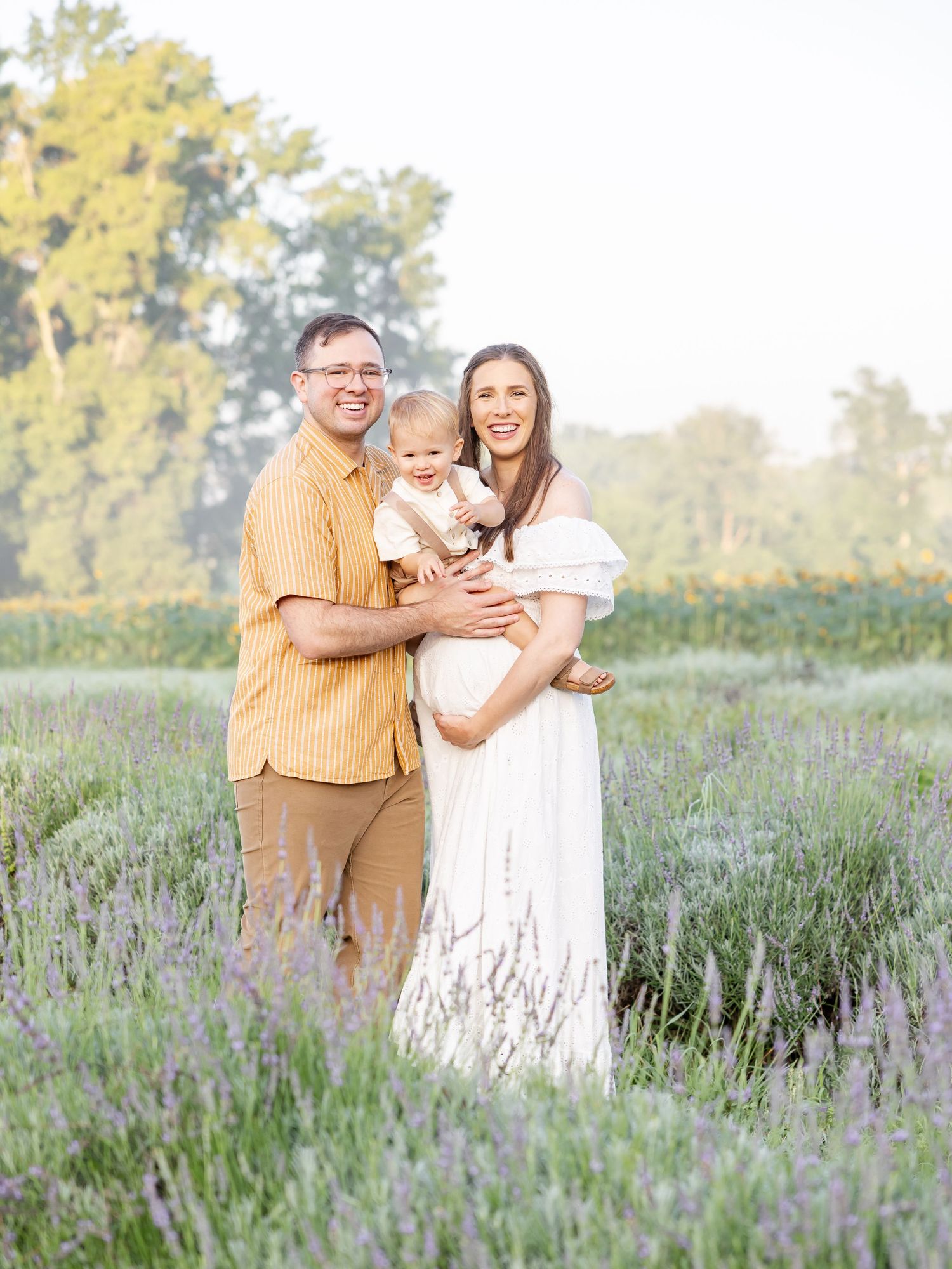 A happy family of three poses in a blooming lavender field, with parents holding their baby on a sunny day.