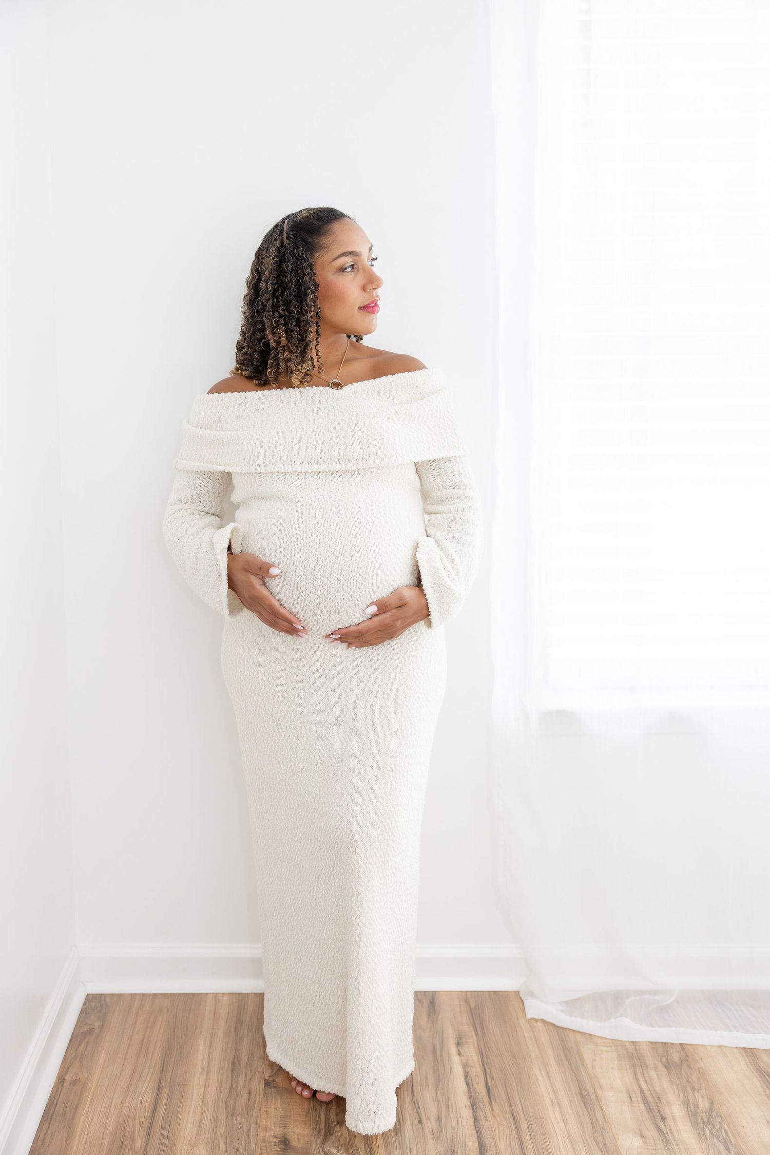 Pregnant woman in white off-shoulder knit maxi dress cradling baby bump, standing against white wall on wood floor.