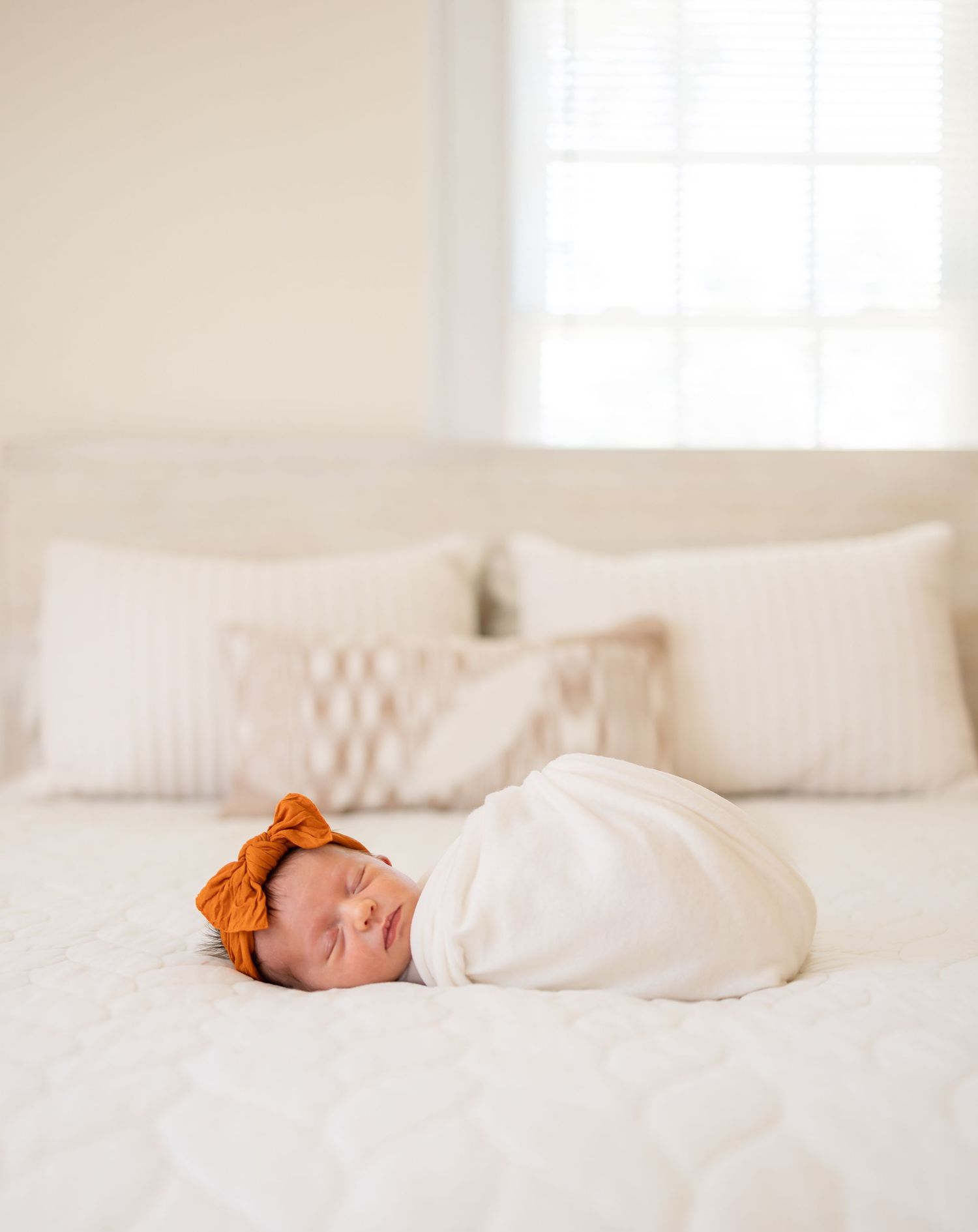 A newborn baby sleeps peacefully swaddled in white blankets with an orange bow headband by Columbus, GA lifestyle newborn photographer.