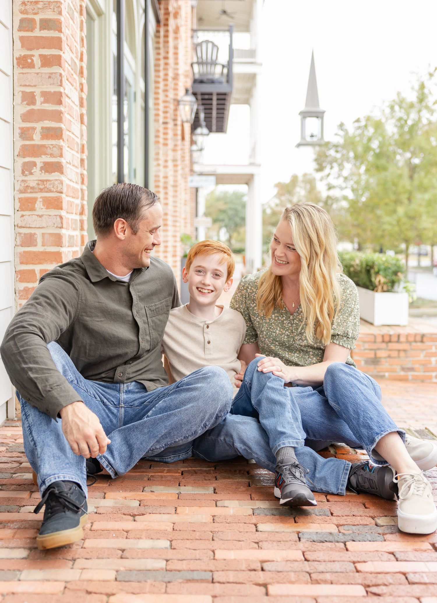 A family sits together smiling and laughing while relaxing on a brick patio in front of a building with a church steeple visible.