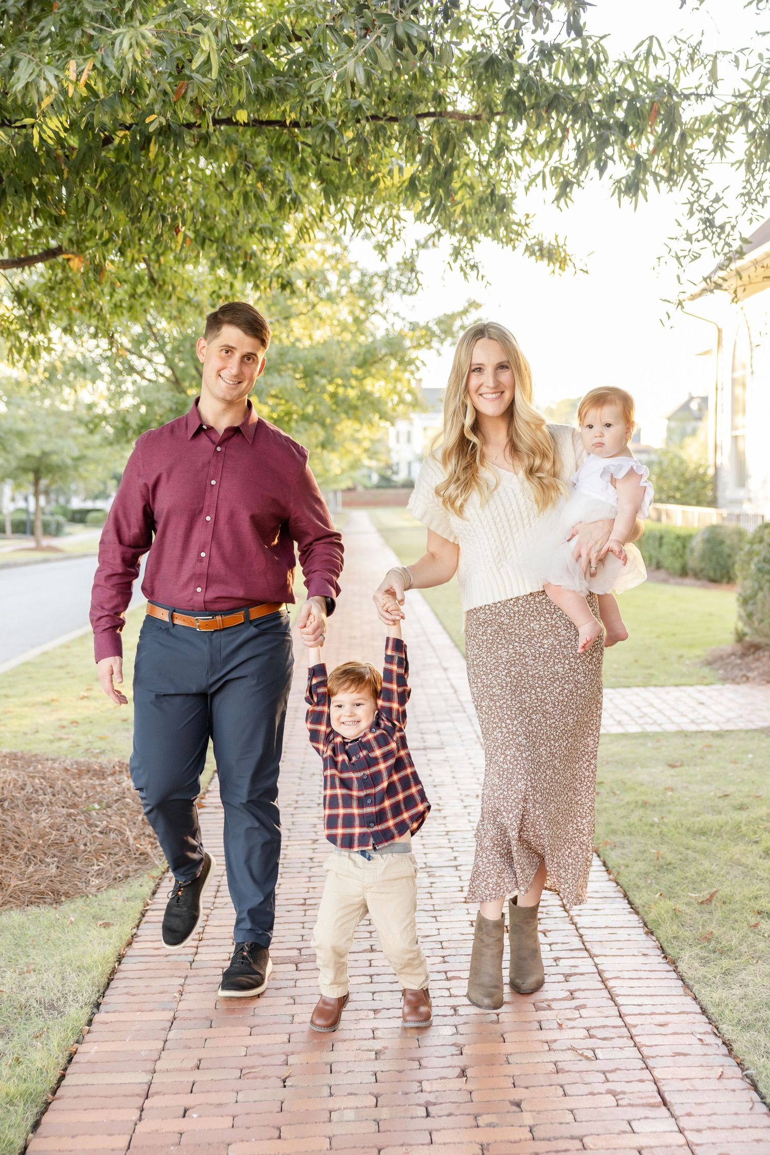 A family of four walks together on a sunlit brick path surrounded by green trees and grass at their Columbus, GA fall portrait session.