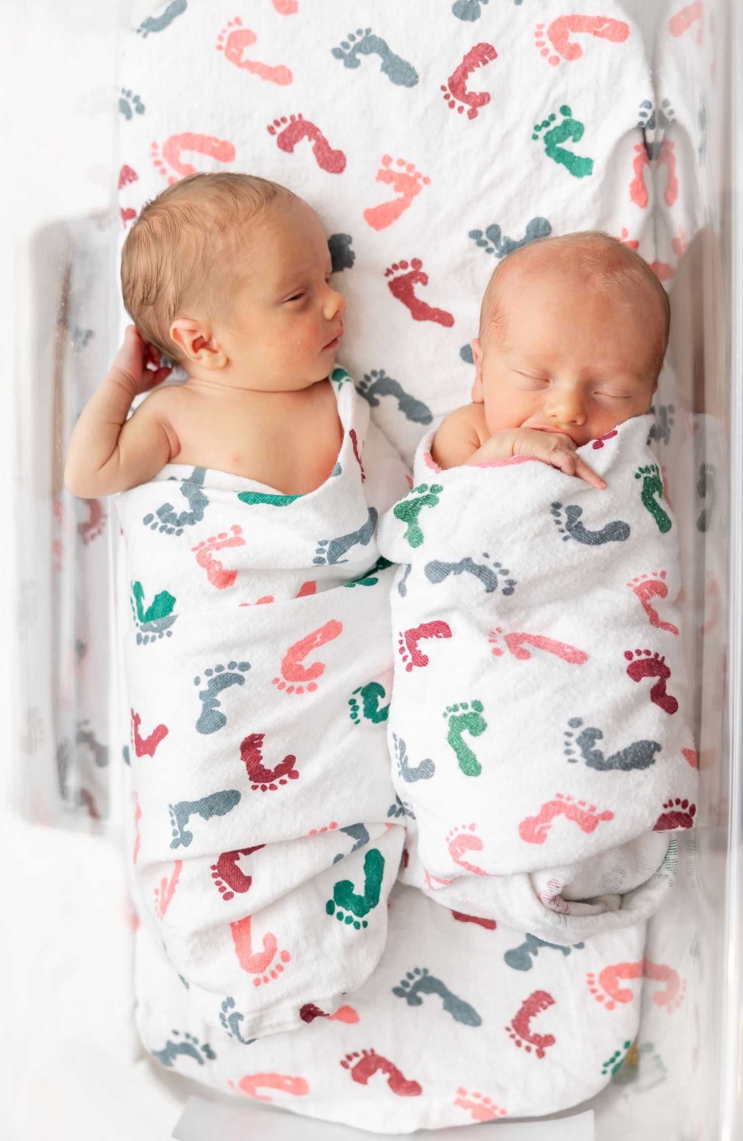 Two swaddled newborns lying together on white fabric with colorful footprint patterns in pink, gray and teal at their Columbus, GA Fresh 48.