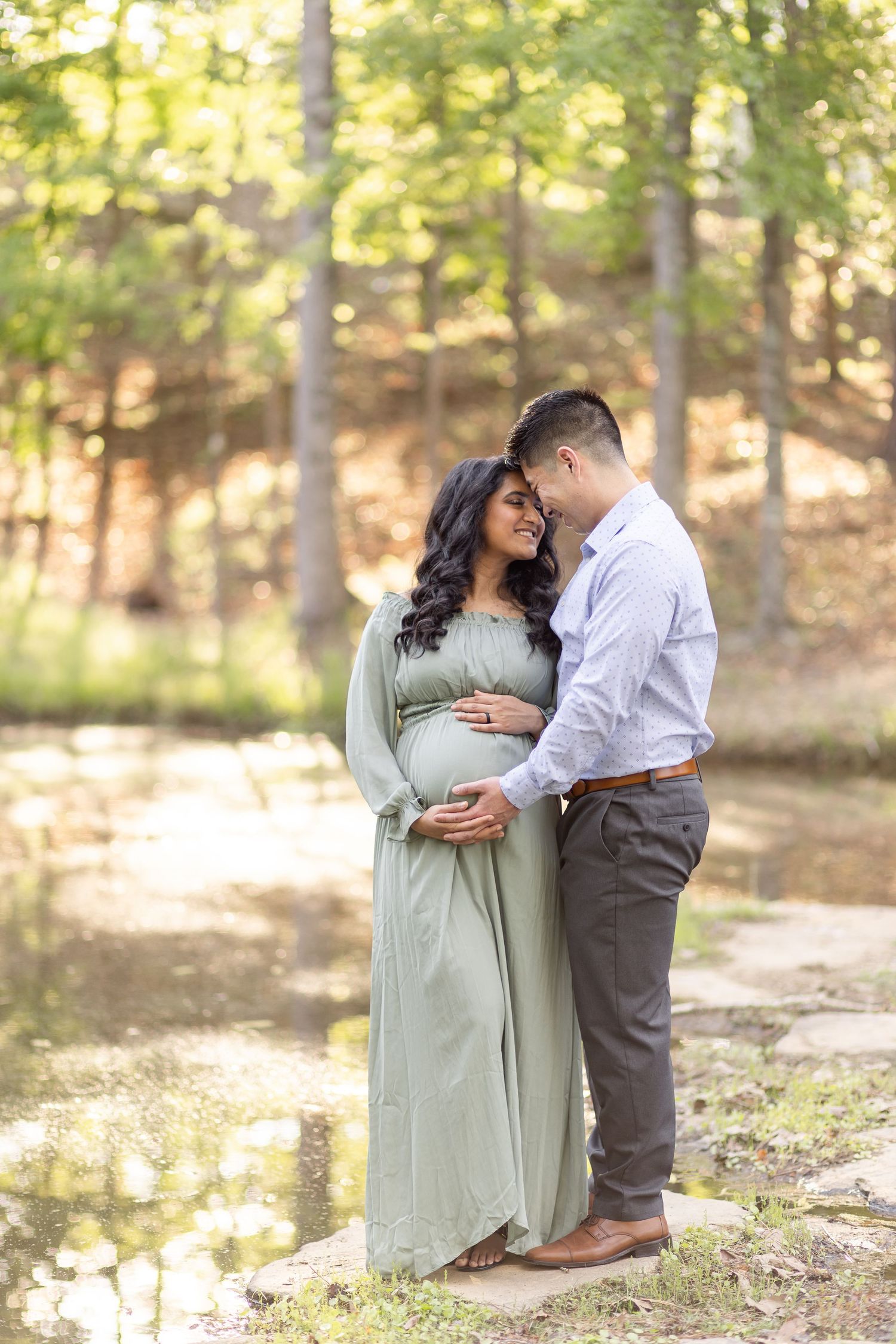 Couple in formal attire shares an intimate moment during outdoor maternity photoshoot in sunlit wooded area in Columbus, GA.