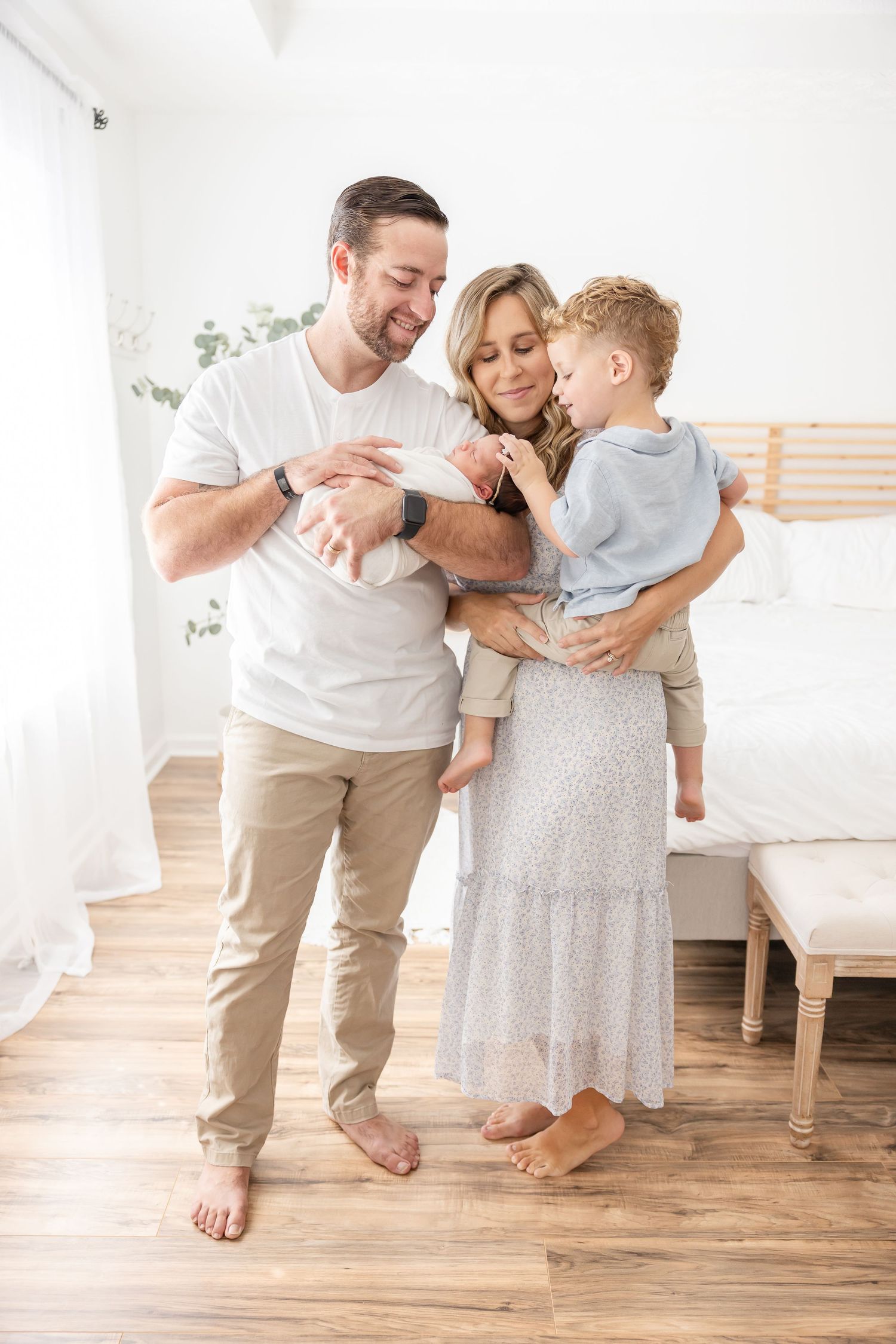 Happy family with parents holding newborn baby and toddler son in bright white bedroom.