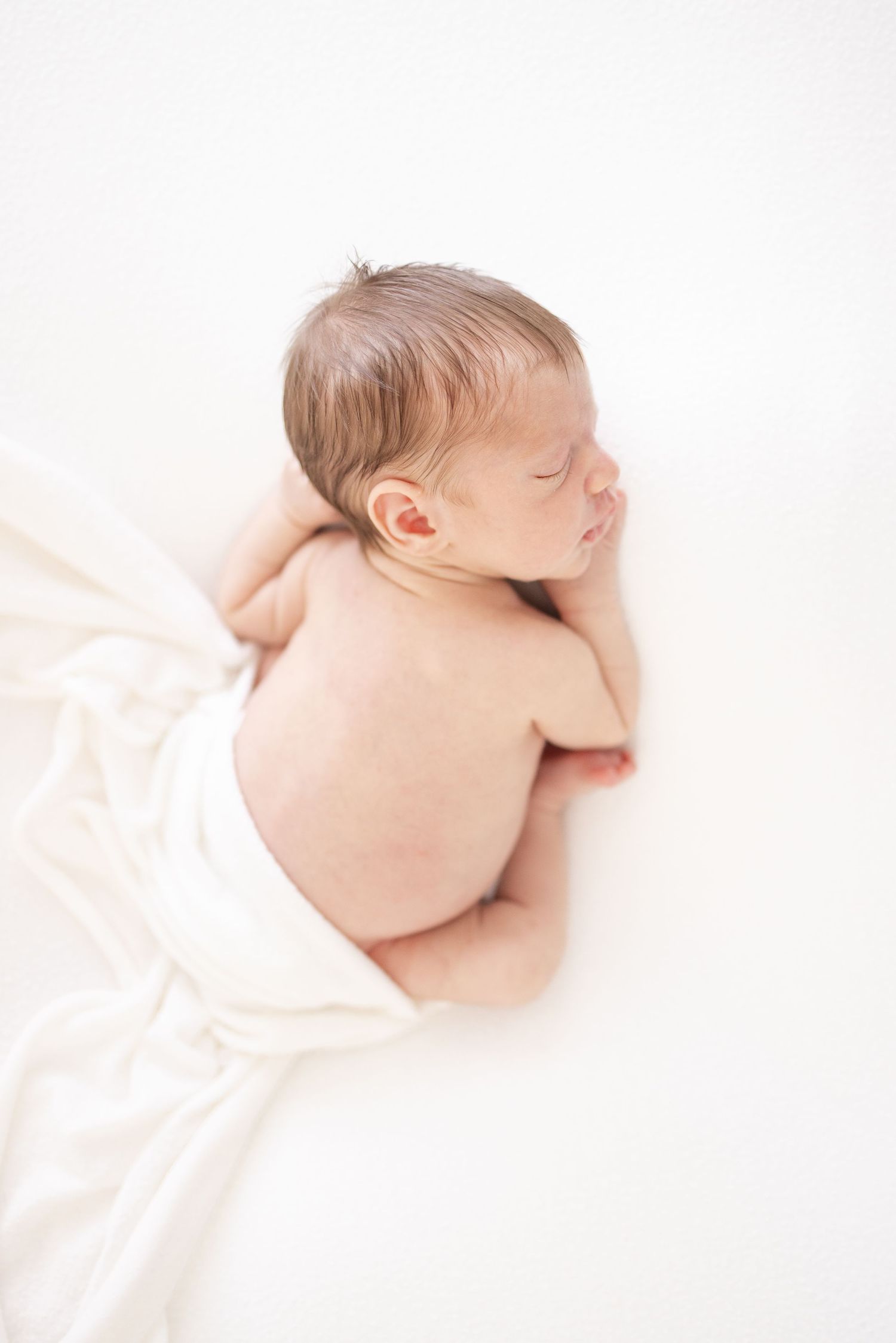 A sleeping newborn baby lies peacefully on soft white fabric in a minimalist portrait style.