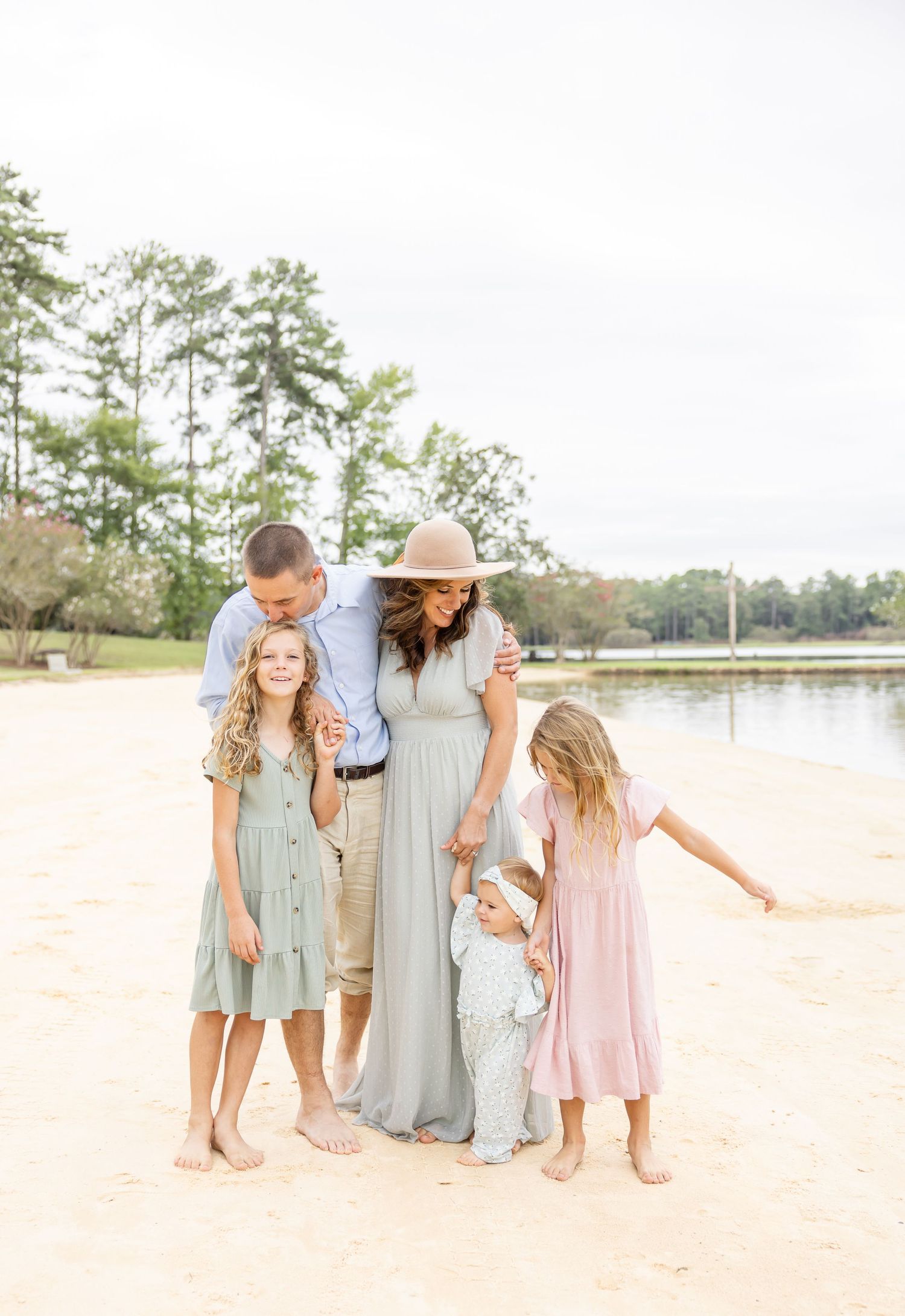 A family in coordinated pastel outfits stands together on a sandy beach near a lake with pine trees in the background.