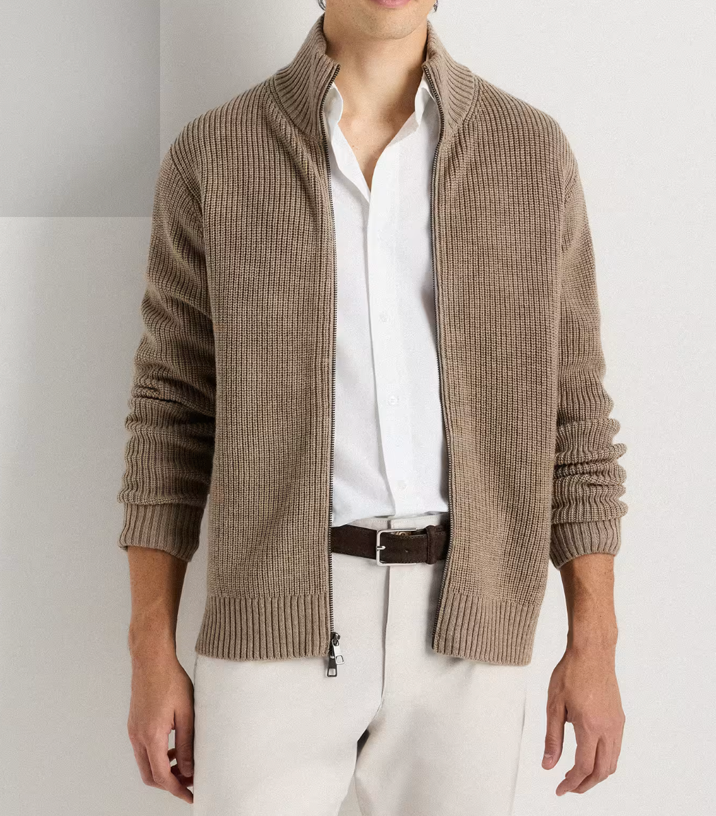 Man wearing a taupe ribbed knit zip-up cardigan over a white shirt with cream trousers and brown belt.