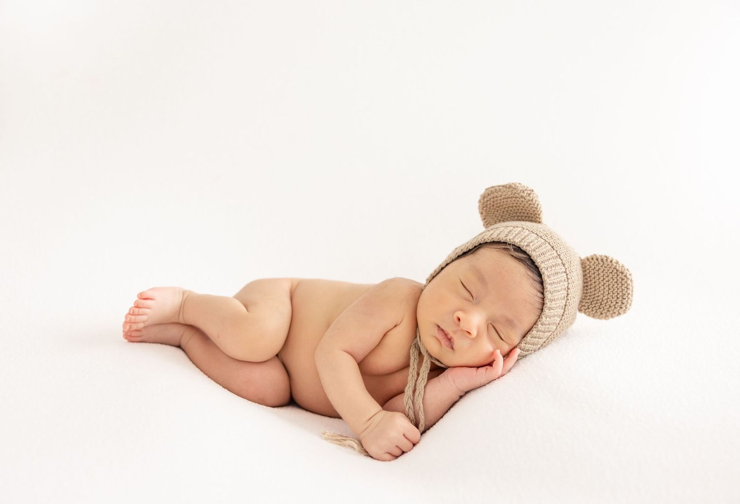 Sleeping newborn baby wearing a knit bear ear bonnet hat, curled up on a white background.