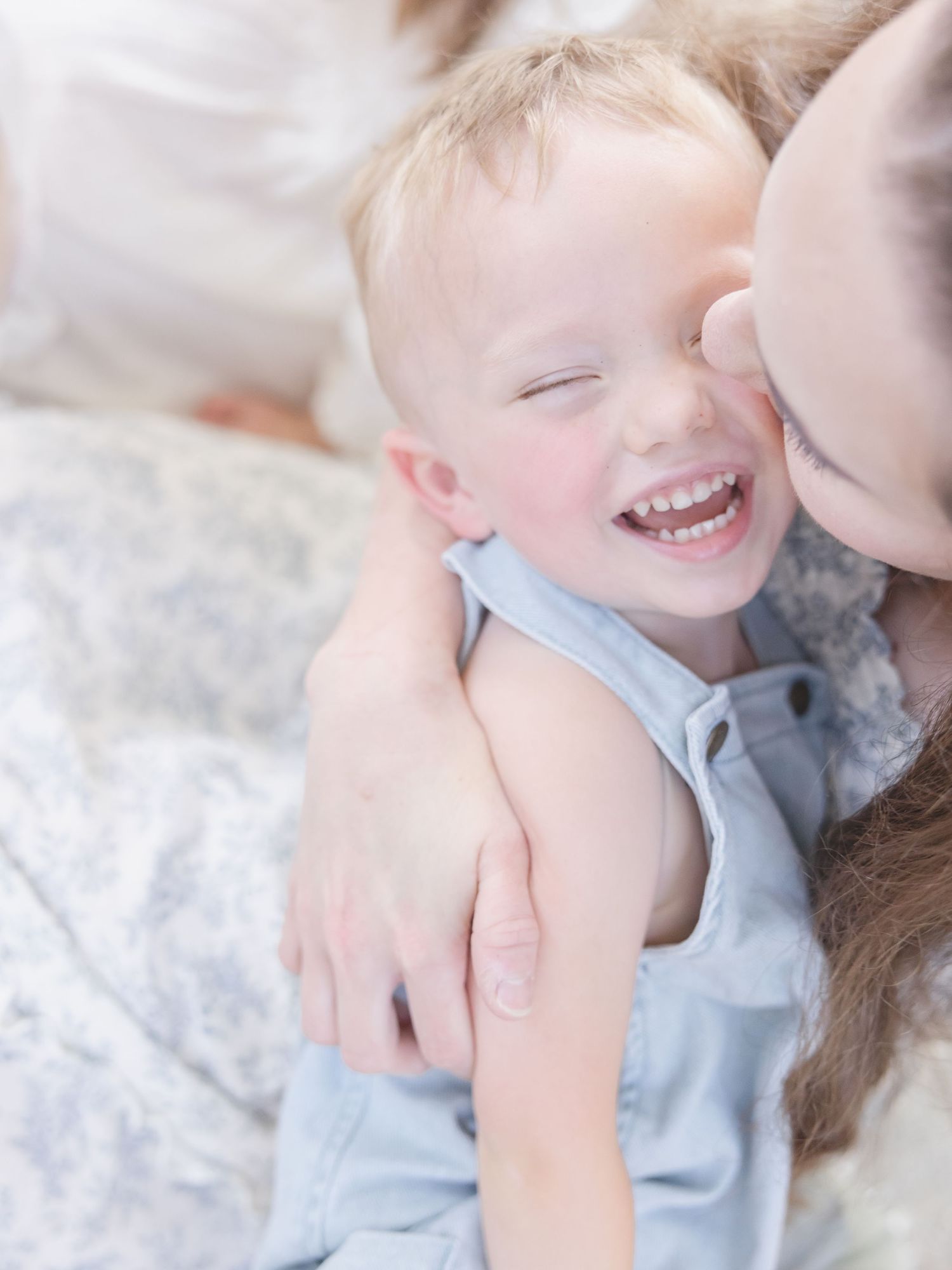 A joyful toddler boy laughs as a woman kisses his cheek, both dressed in soft blue tones in a bright, airy setting.