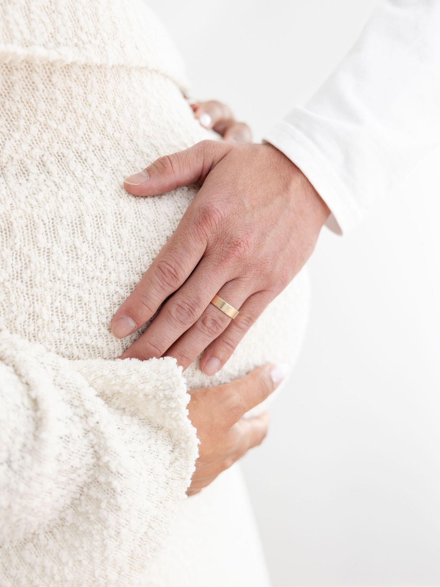 A partner's hand with a gold wedding ring resting on a pregnant woman's baby bump in a white sweater.