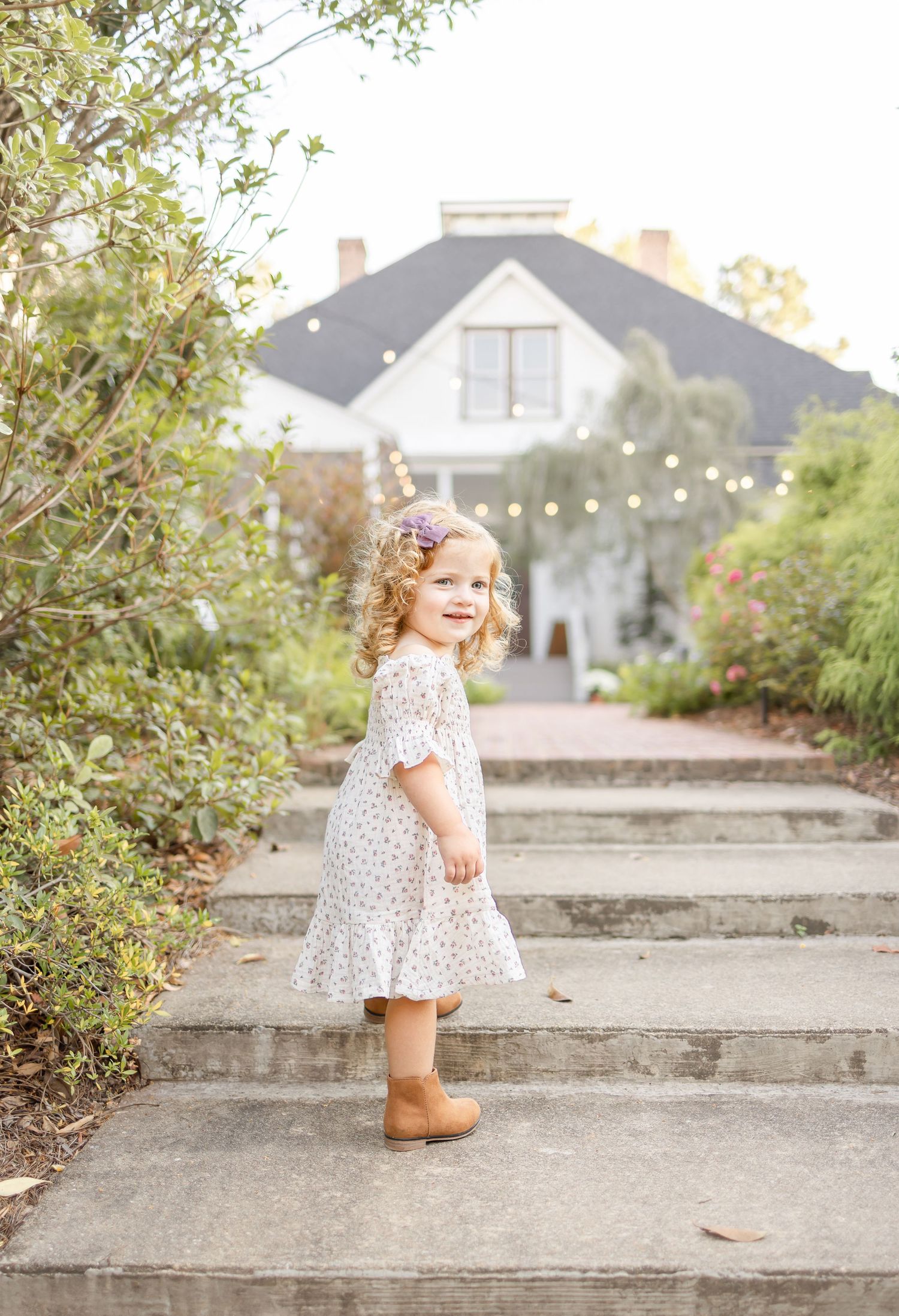 Small child wearing a floral dress and brown boots stands on concrete steps outside a large gray house.