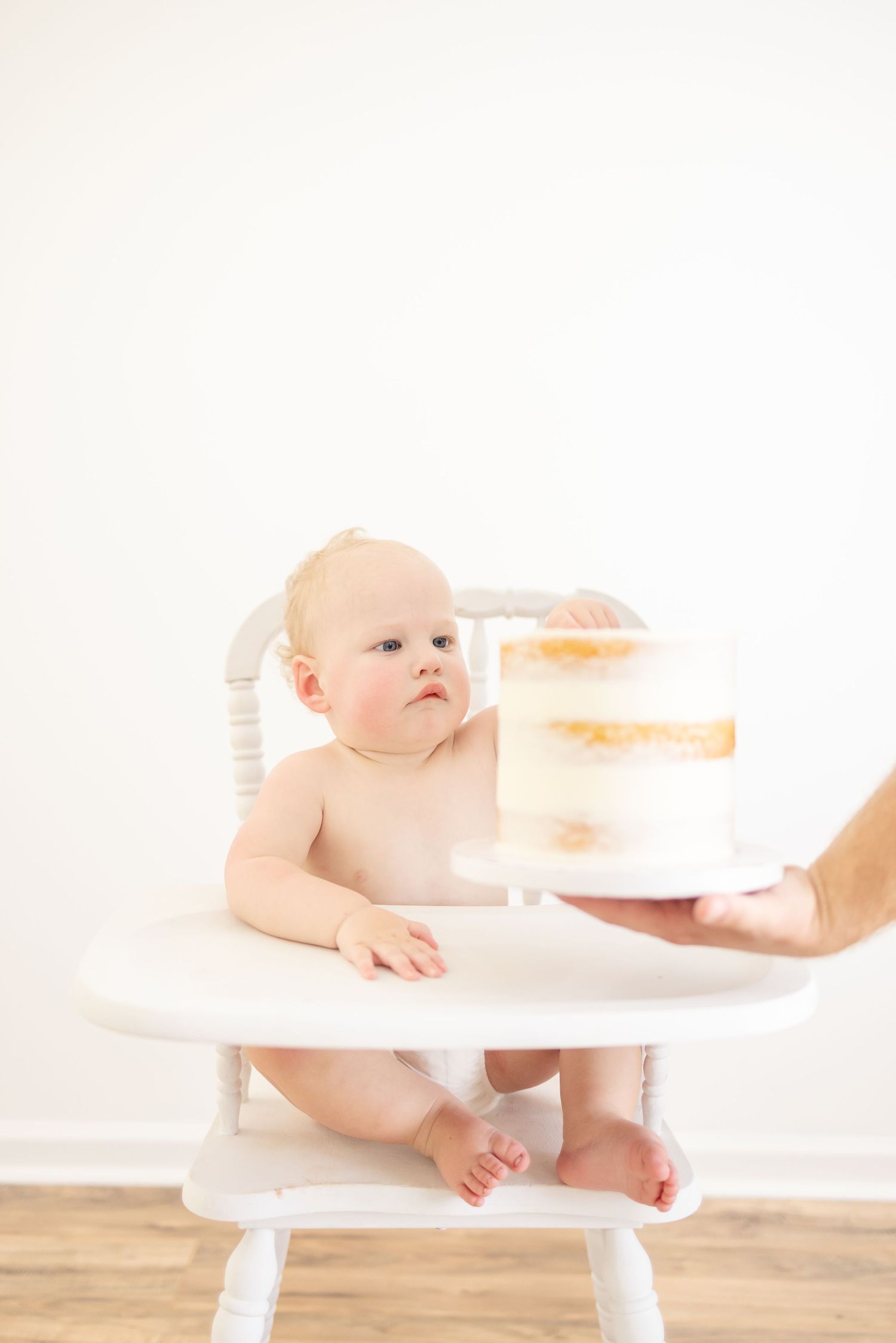 Baby seated in white high chair being present with a naked white cake at his first birthday photoshoot.