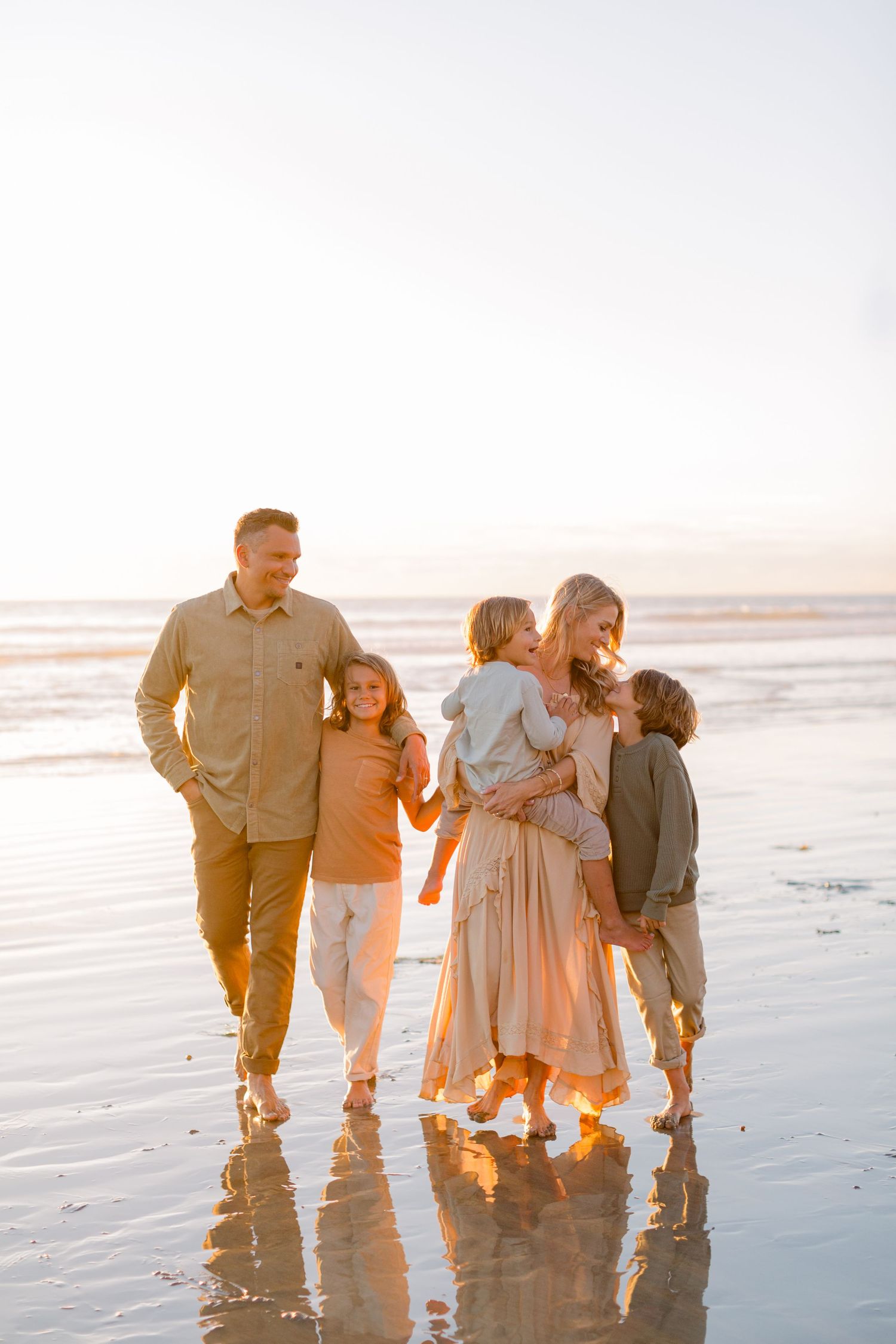 Family of five walking on a reflective beach at sunset, smiling and embracing in warm neutral tones.