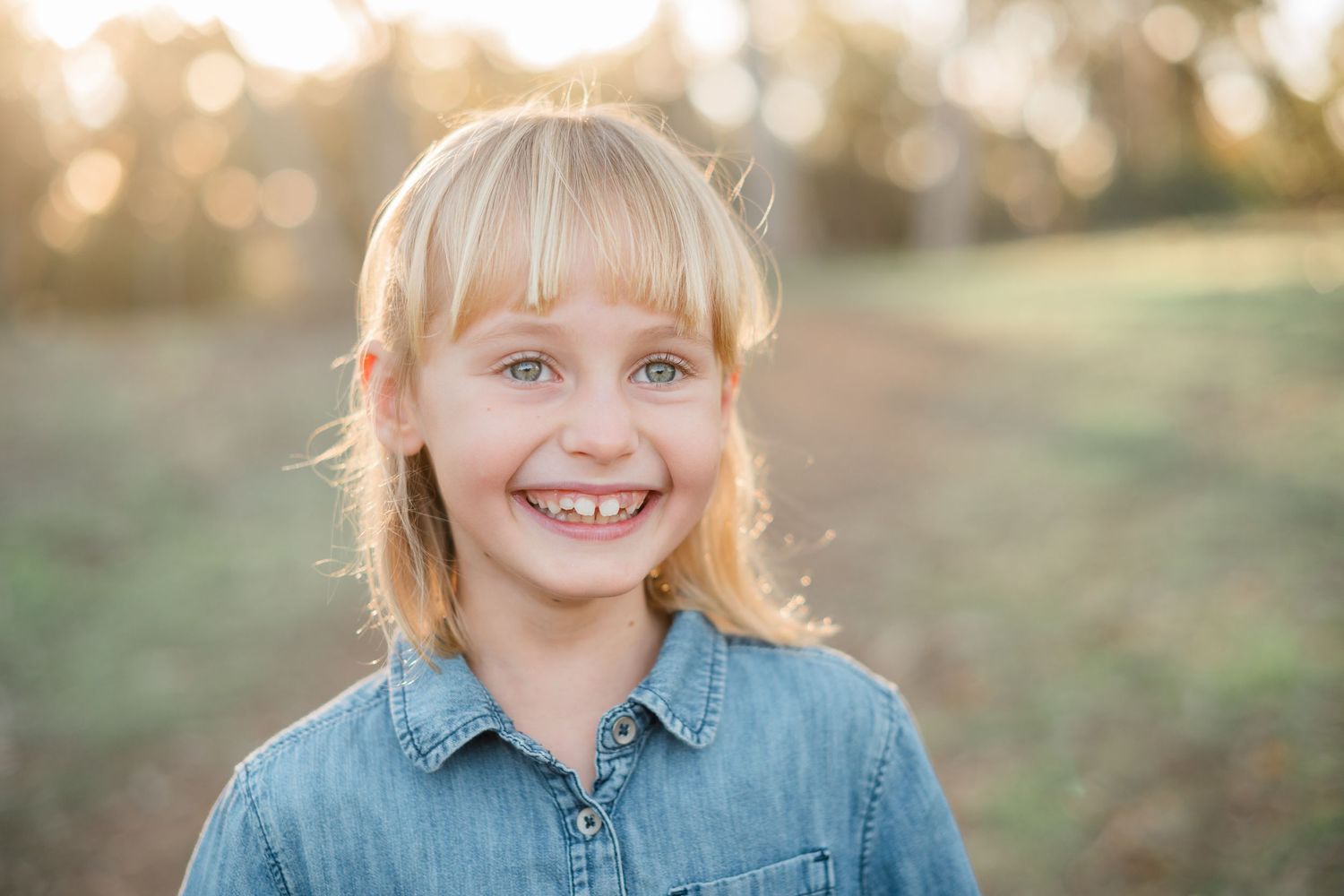 Smiling blonde girl with blue eyes wearing a denim shirt outdoors in a sunny field.