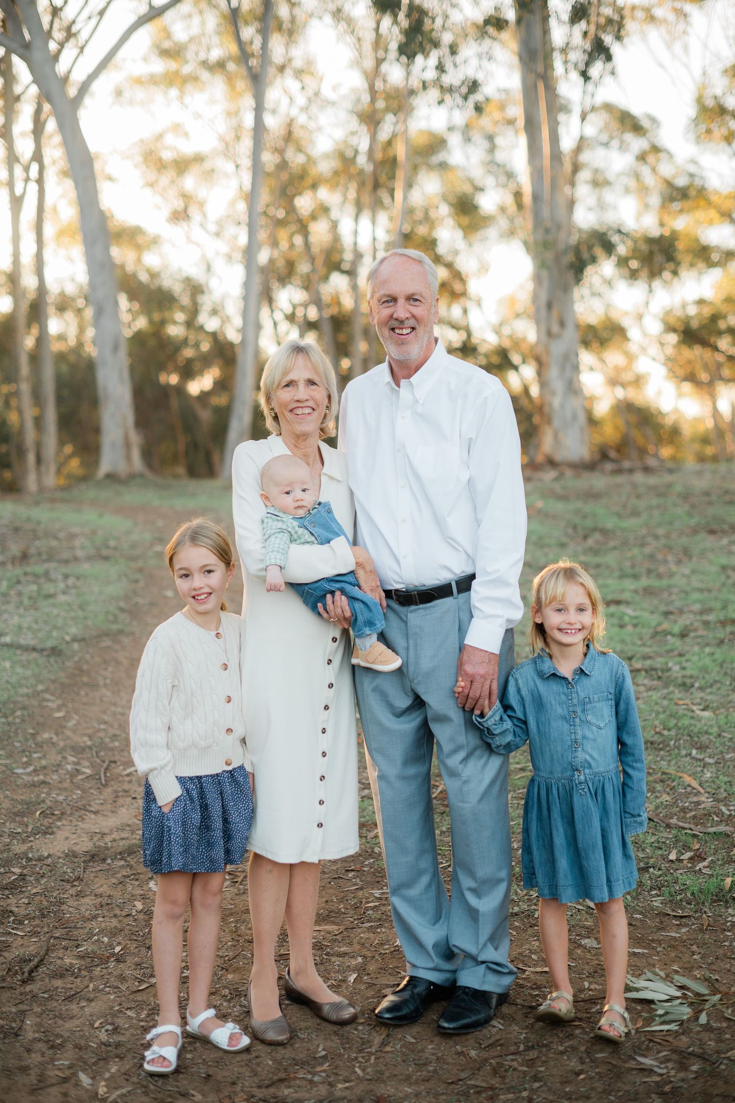 Grandparents pose outdoors with three young grandchildren, including a baby, surrounded by tall trees.