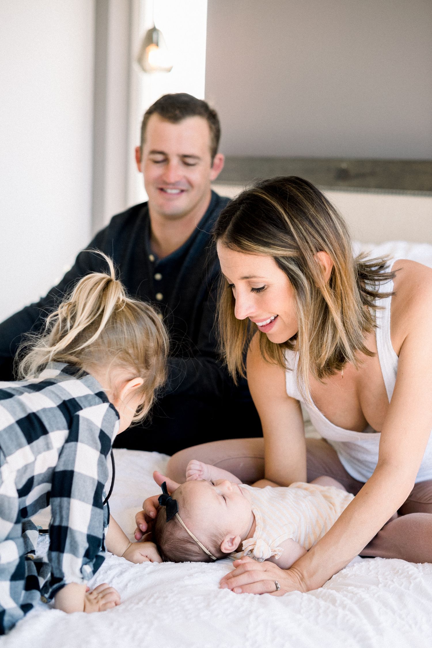 Smiling parents and toddler gathered around their newborn baby lying on a white bed at home.