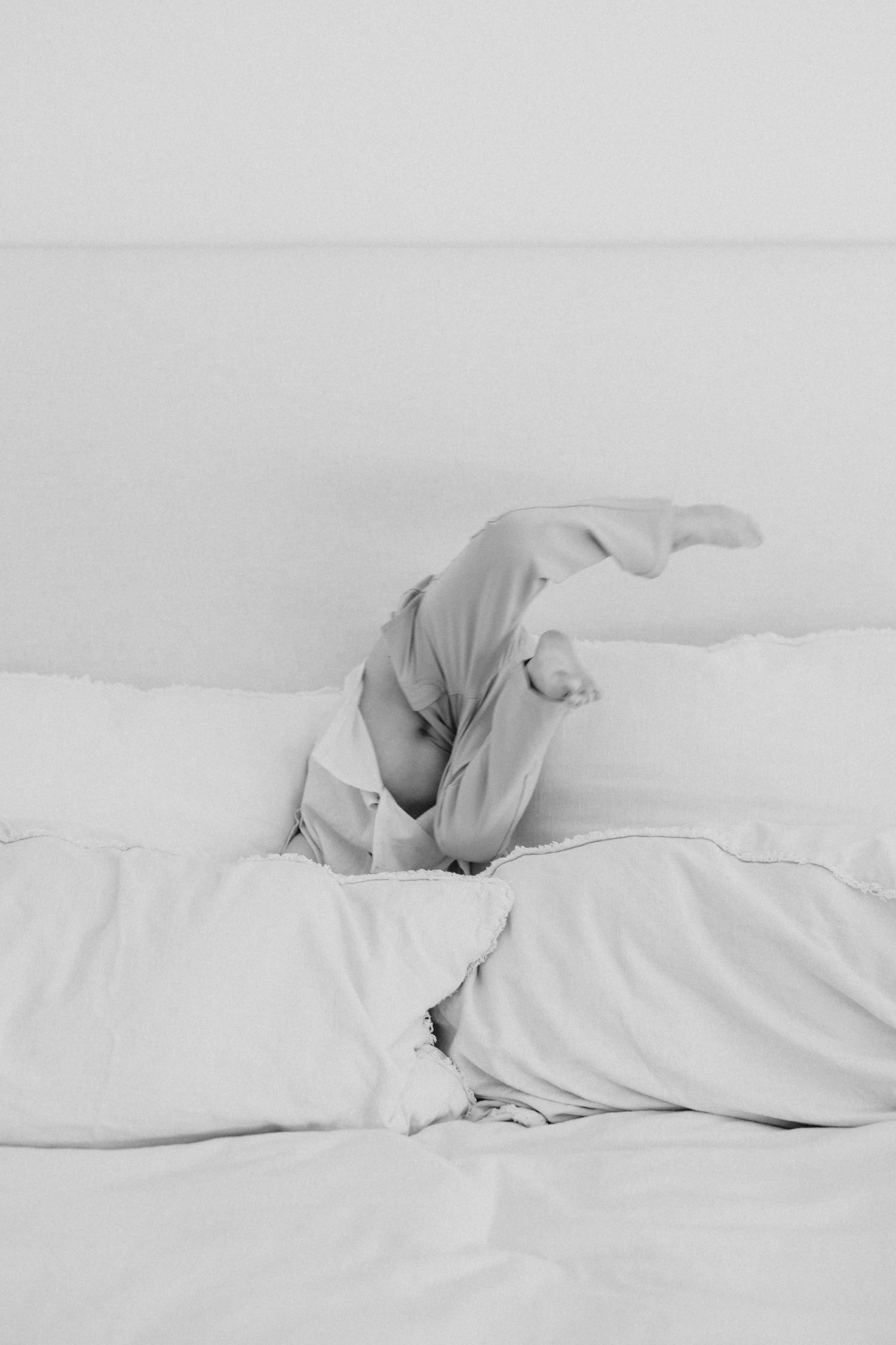 Black and white photo of person lying upside down on white bed with legs up, surrounded by soft pillows.