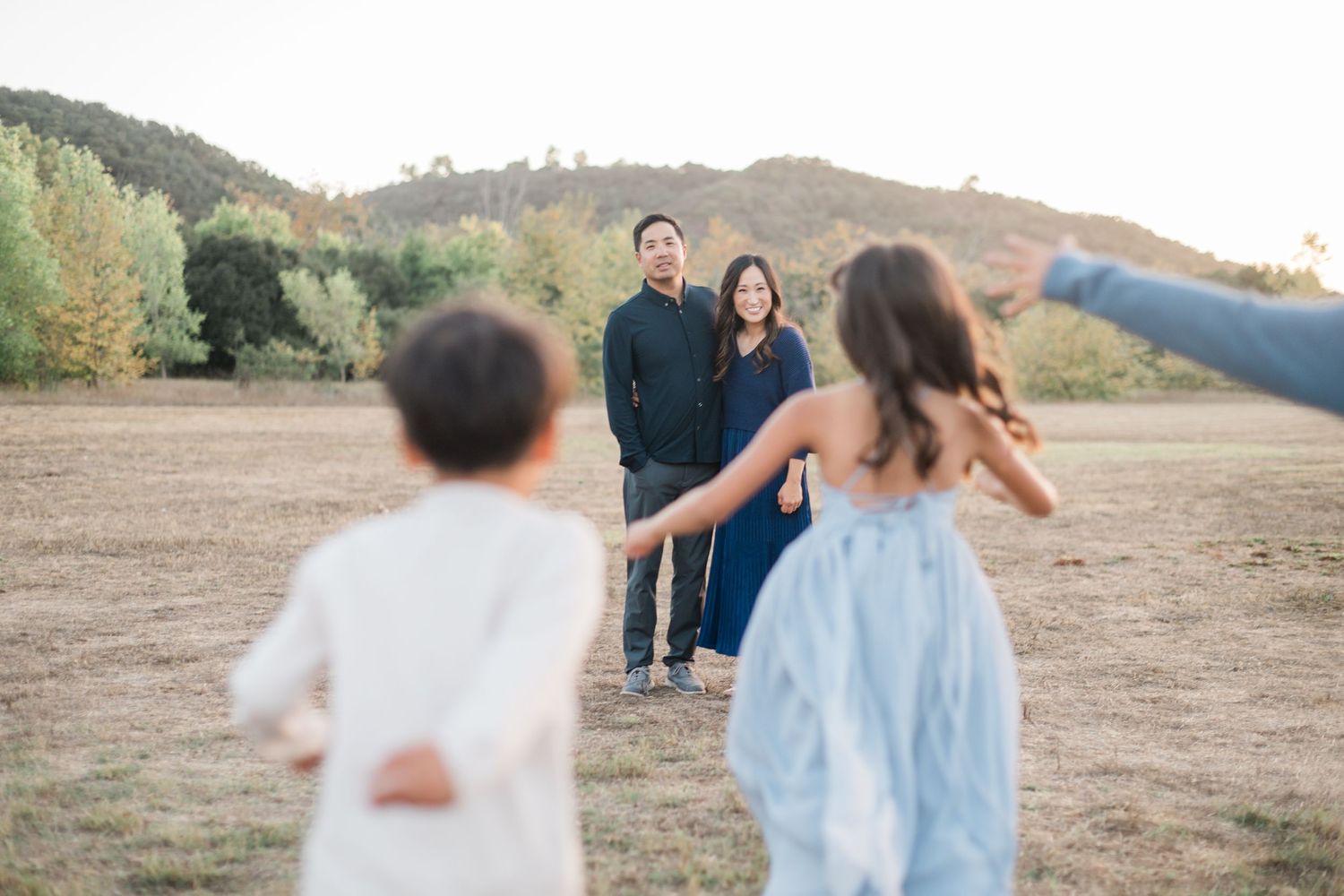 Family portrait session in a golden field with parents standing together as two children run toward them at sunset.