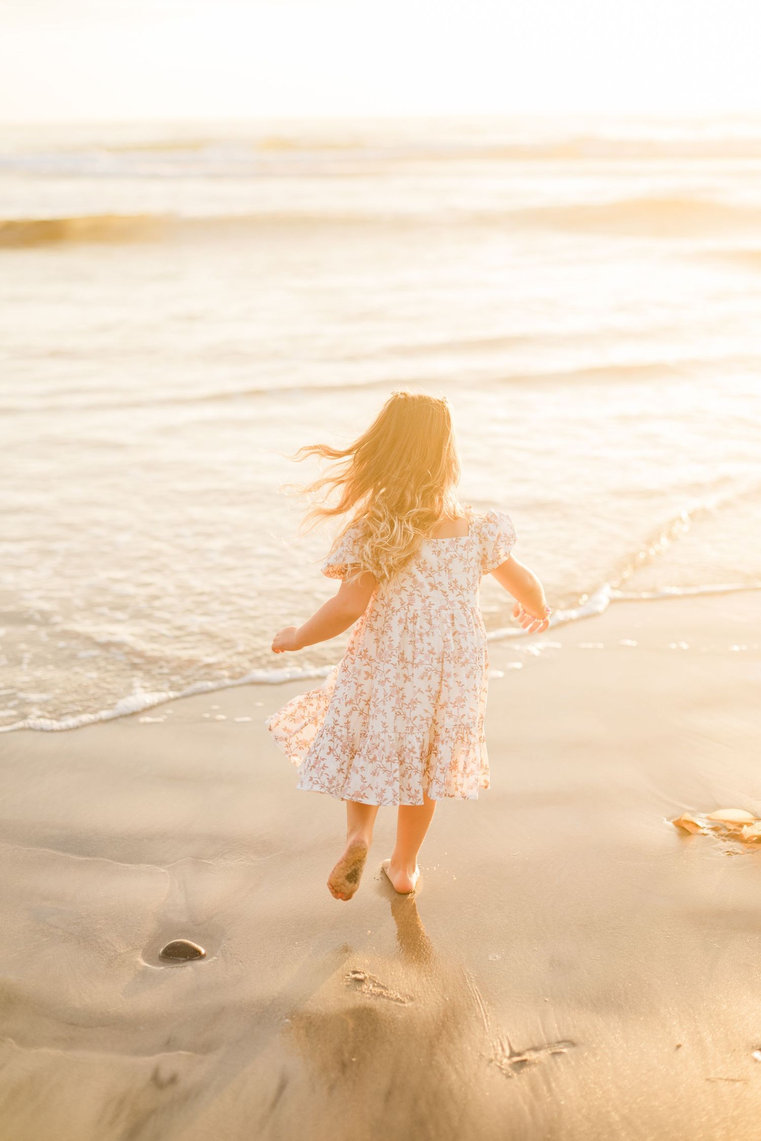 Young girl in floral dress running toward ocean waves on sandy beach at golden sunset.