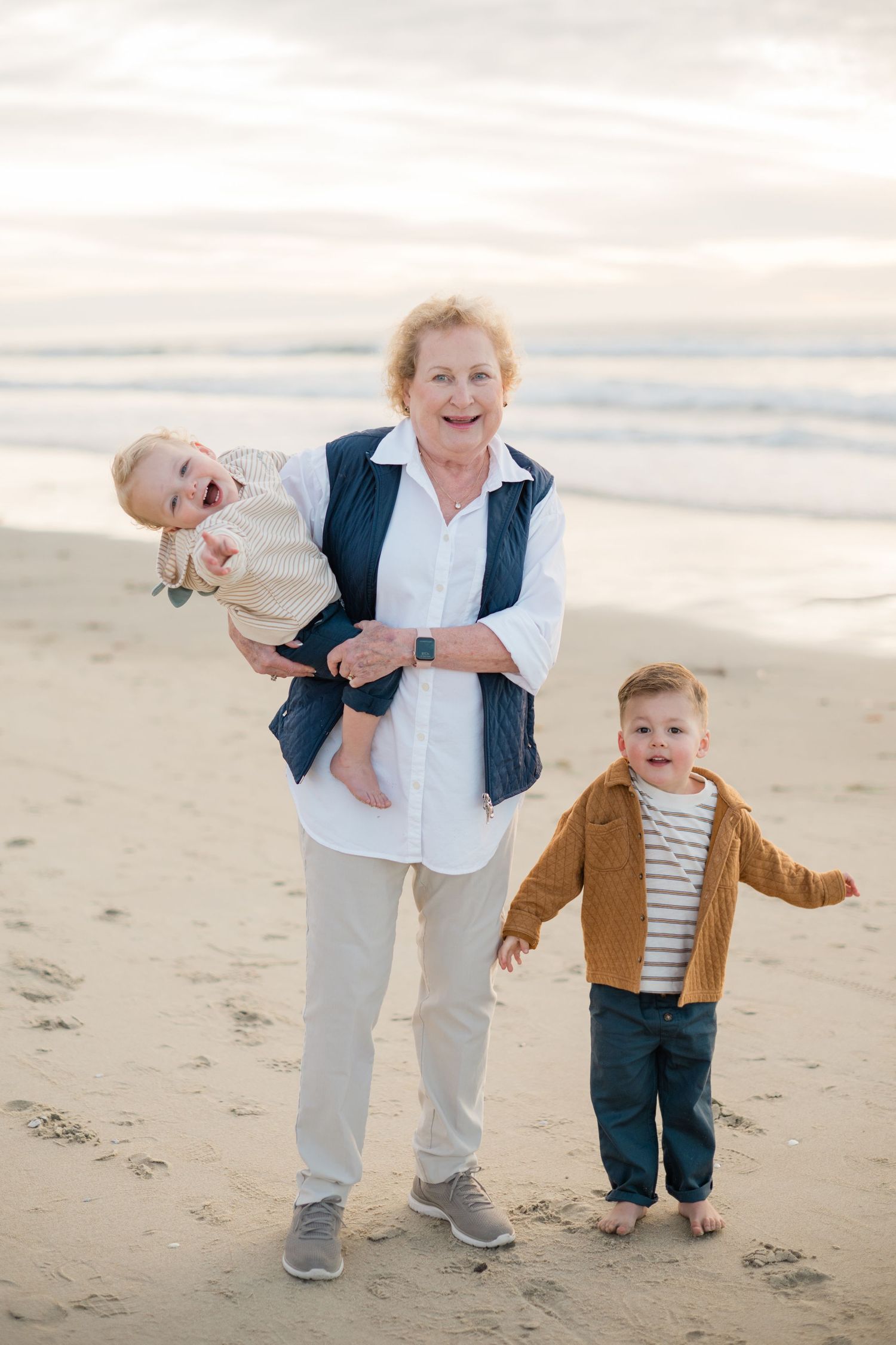 Grandmother holds baby while toddler stands beside her on a sandy beach at sunset.