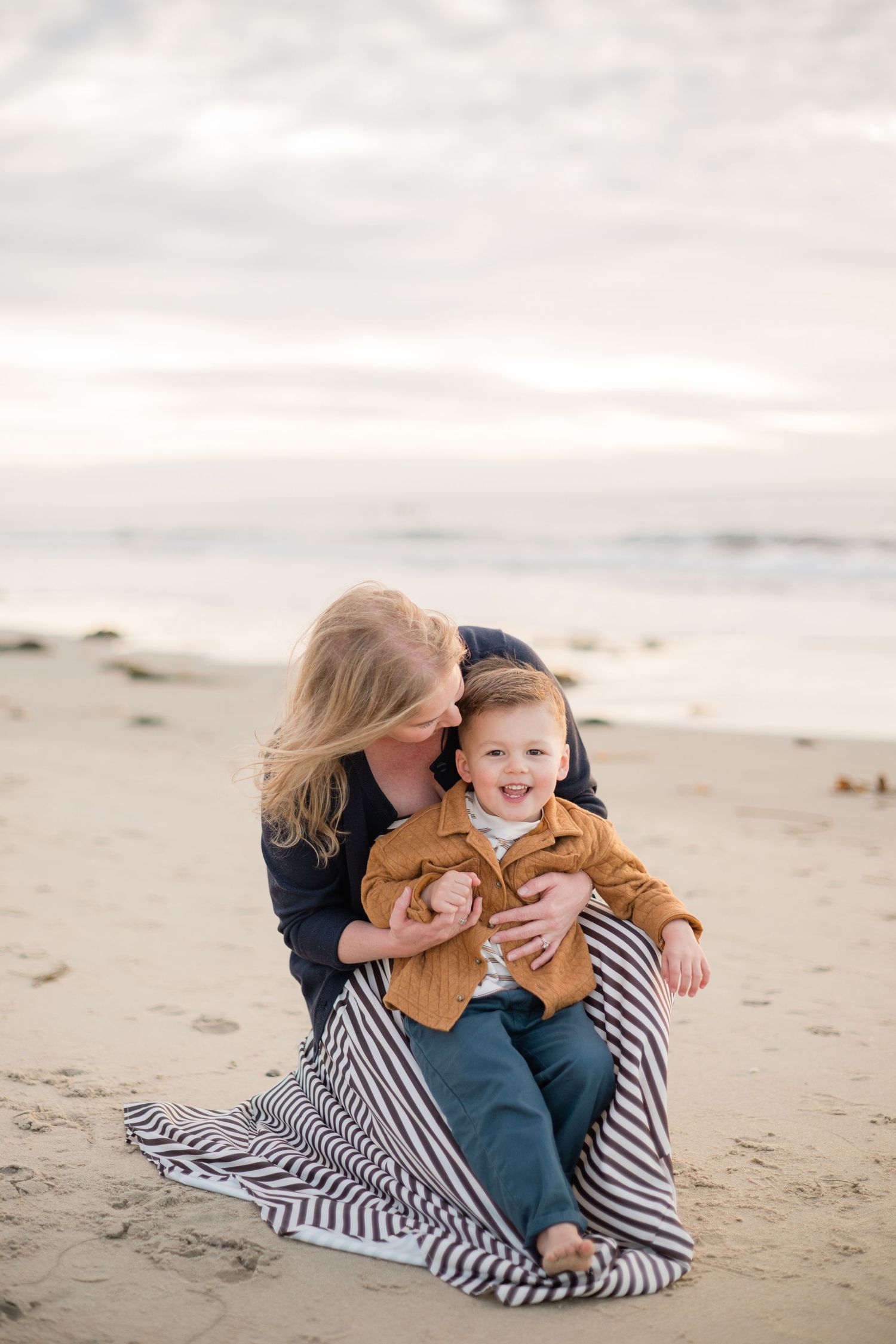 Blonde mother sitting on sandy beach kissing smiling toddler boy in brown jacket at golden hour.