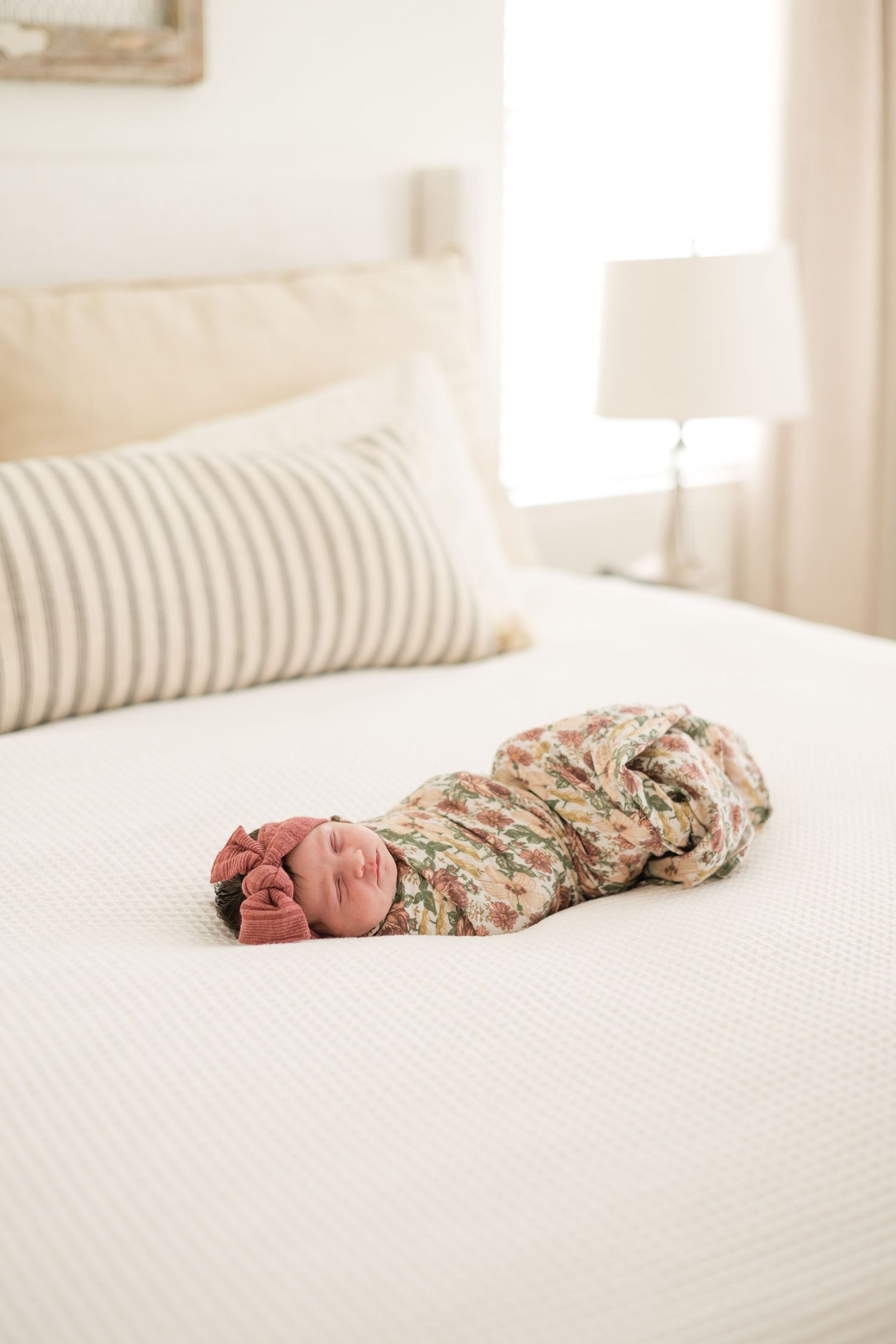 Swaddled newborn baby in floral wrap with pink bow lying on white bed with striped pillow.