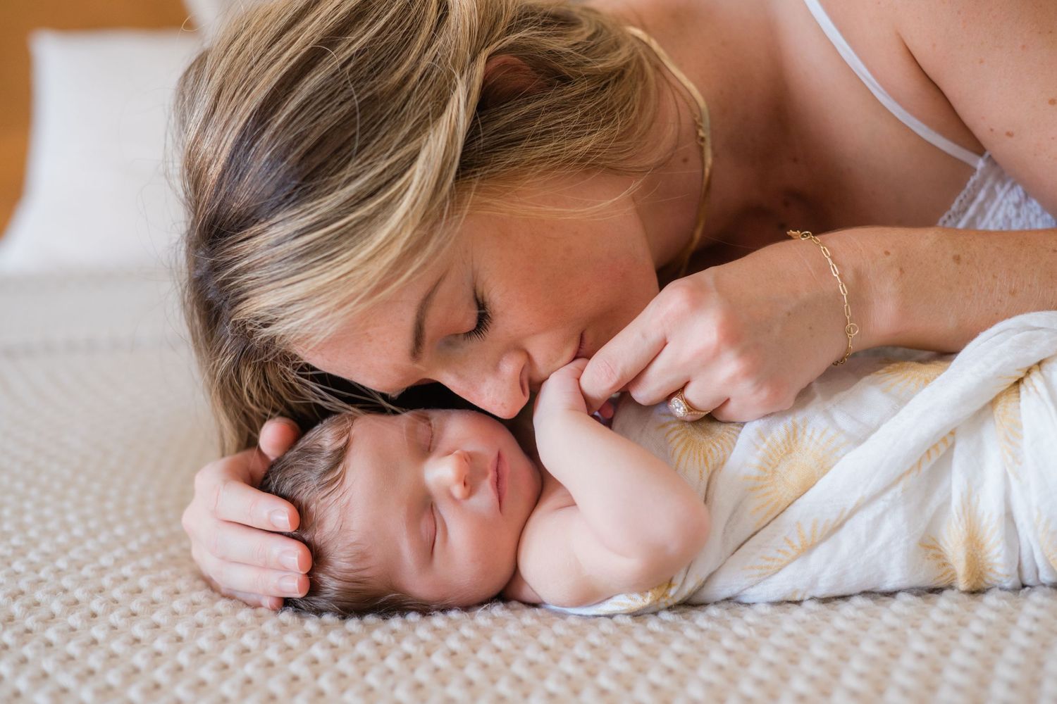 A mother tenderly kisses her sleeping newborn baby swaddled in a white blanket on a bed.
