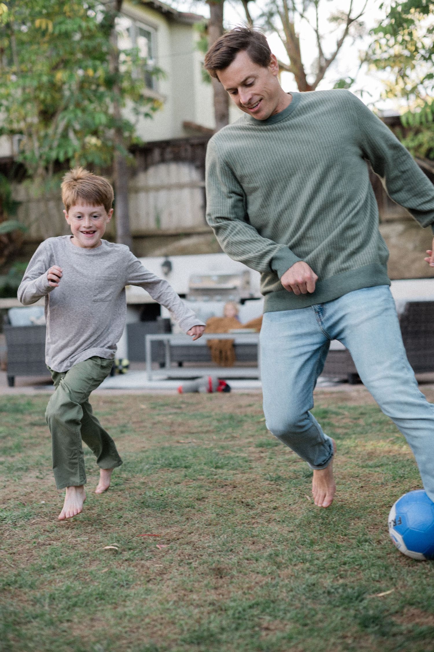 A father and son play soccer together barefoot in a backyard lawn.