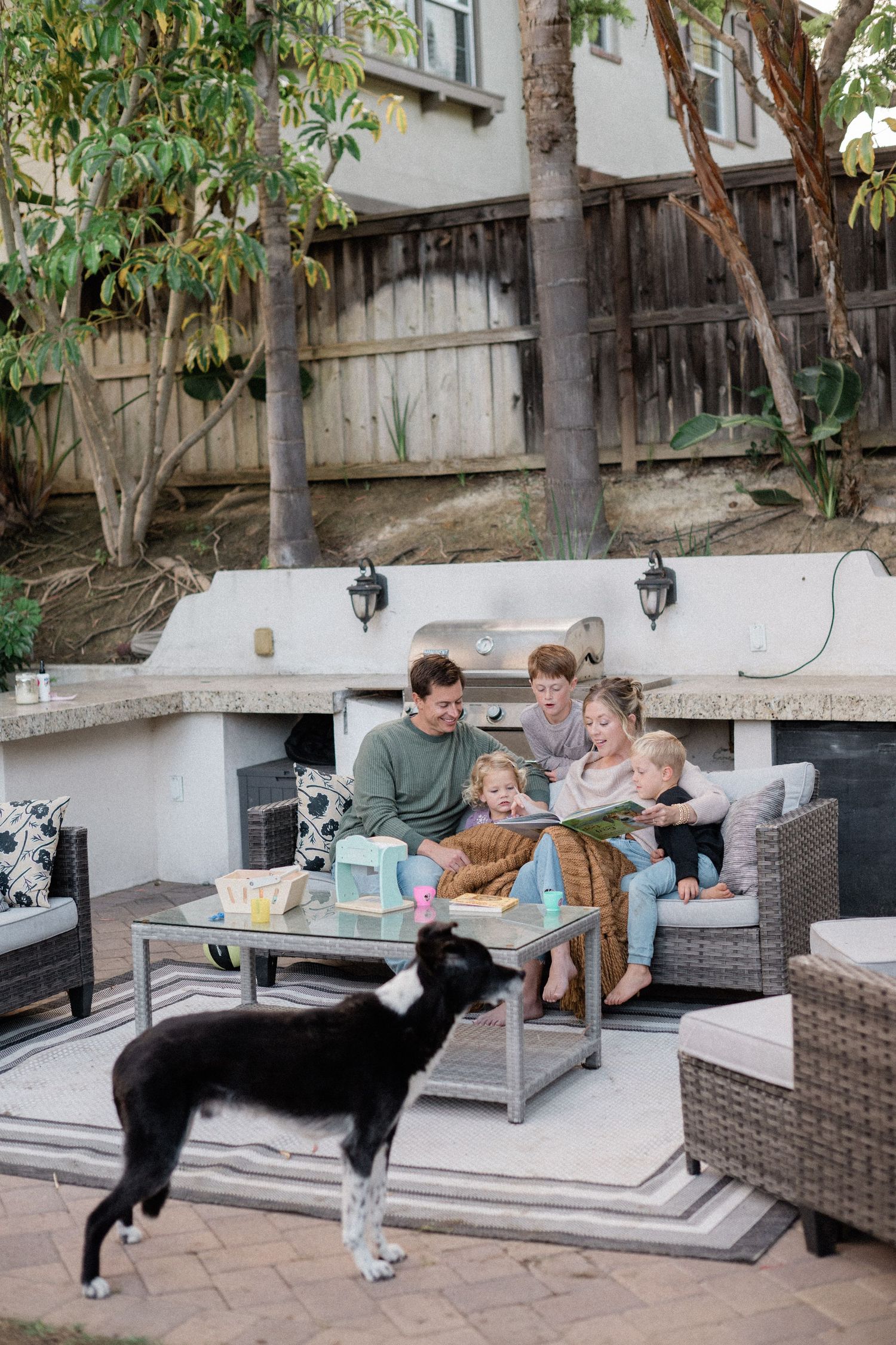 Family reading together on patio furniture in backyard with black and white dog in foreground.
