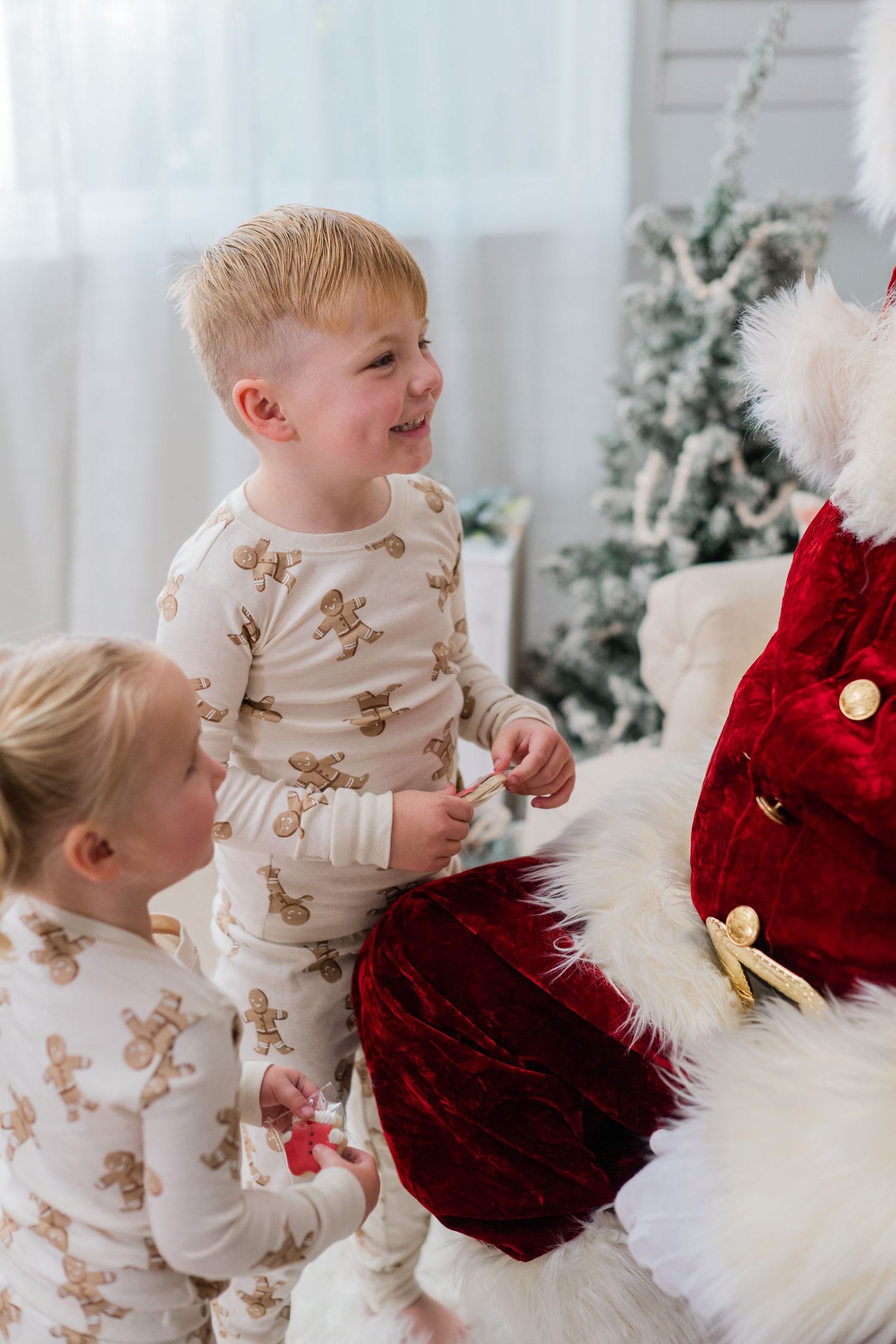 Two young children in gingerbread pajamas smile while standing next to Santa Claus near a Christmas tree.