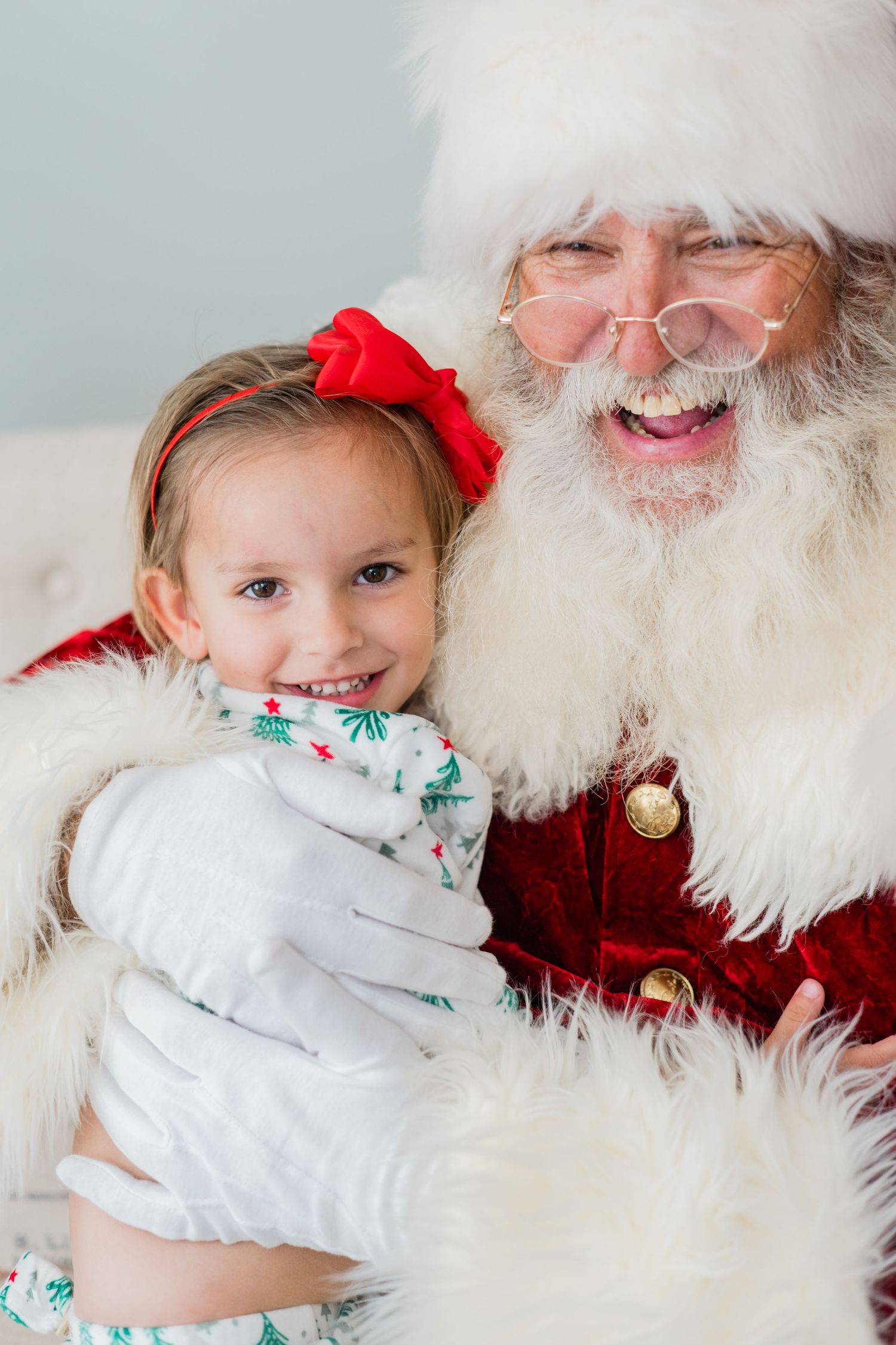 Happy little girl with red bow smiling while being hugged by Santa Claus in a Christmas portrait.