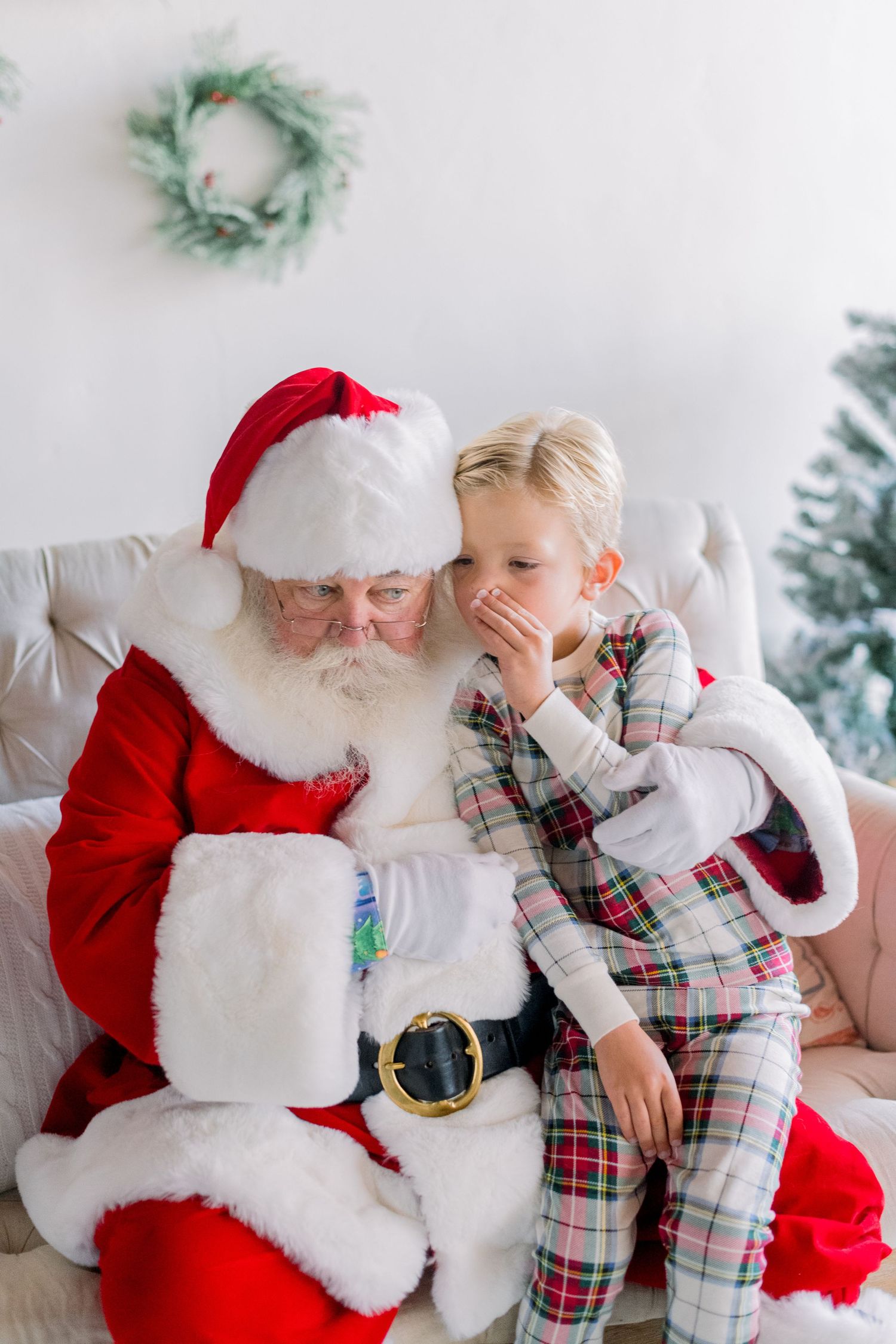 A blonde child in plaid pajamas whispers a Christmas wish into Santa Claus's ear while sitting on a couch.