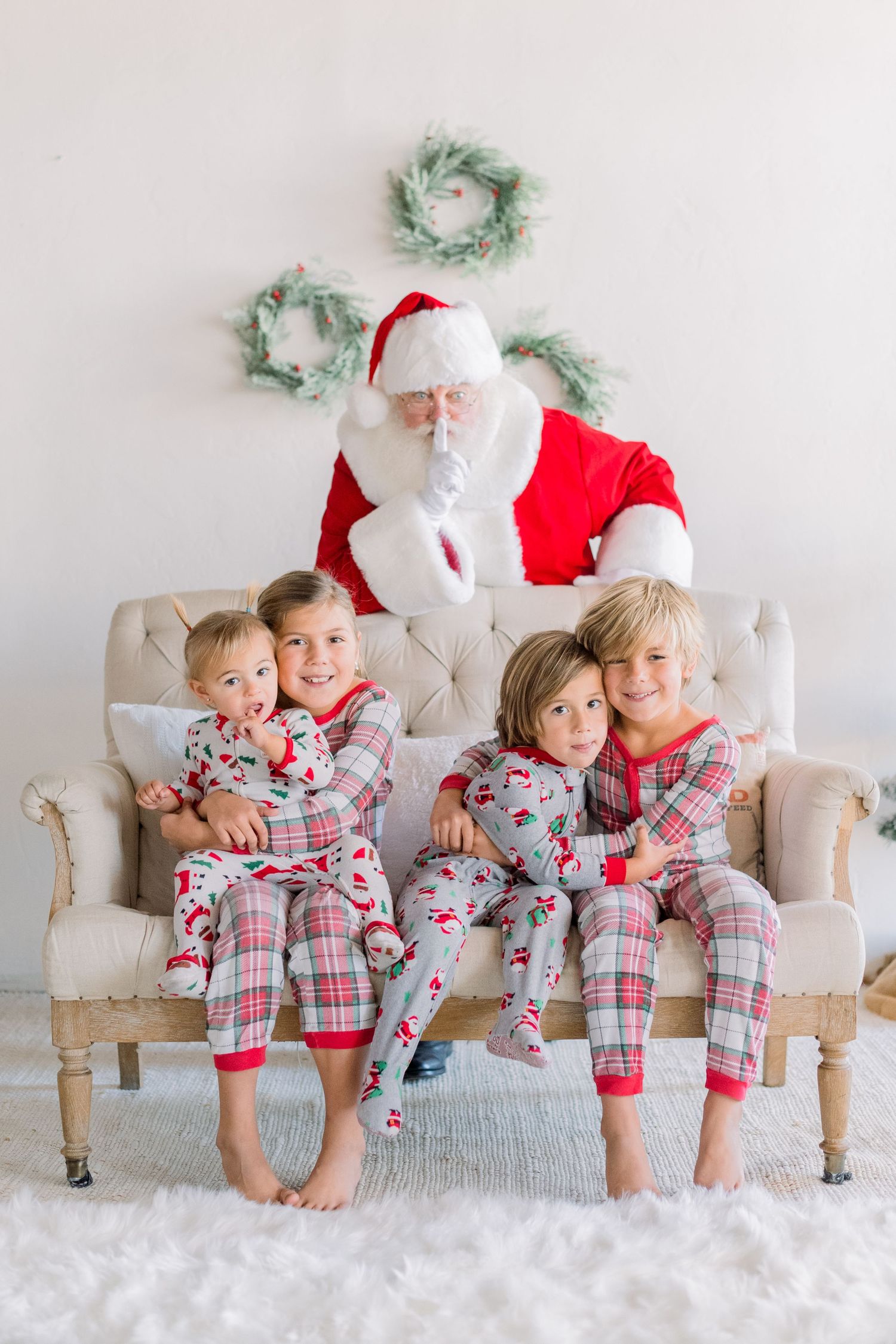 Four children in matching Christmas pajamas pose with Santa Claus on a cozy chair in a festive holiday setting.