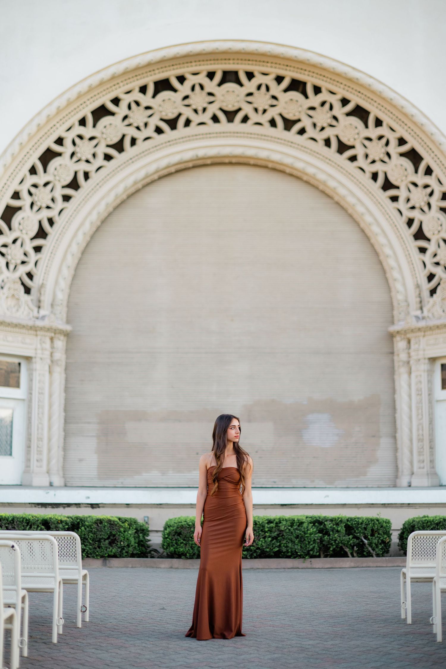 Woman in elegant brown maxi dress poses before ornate arched architectural detail at Balboa Park.