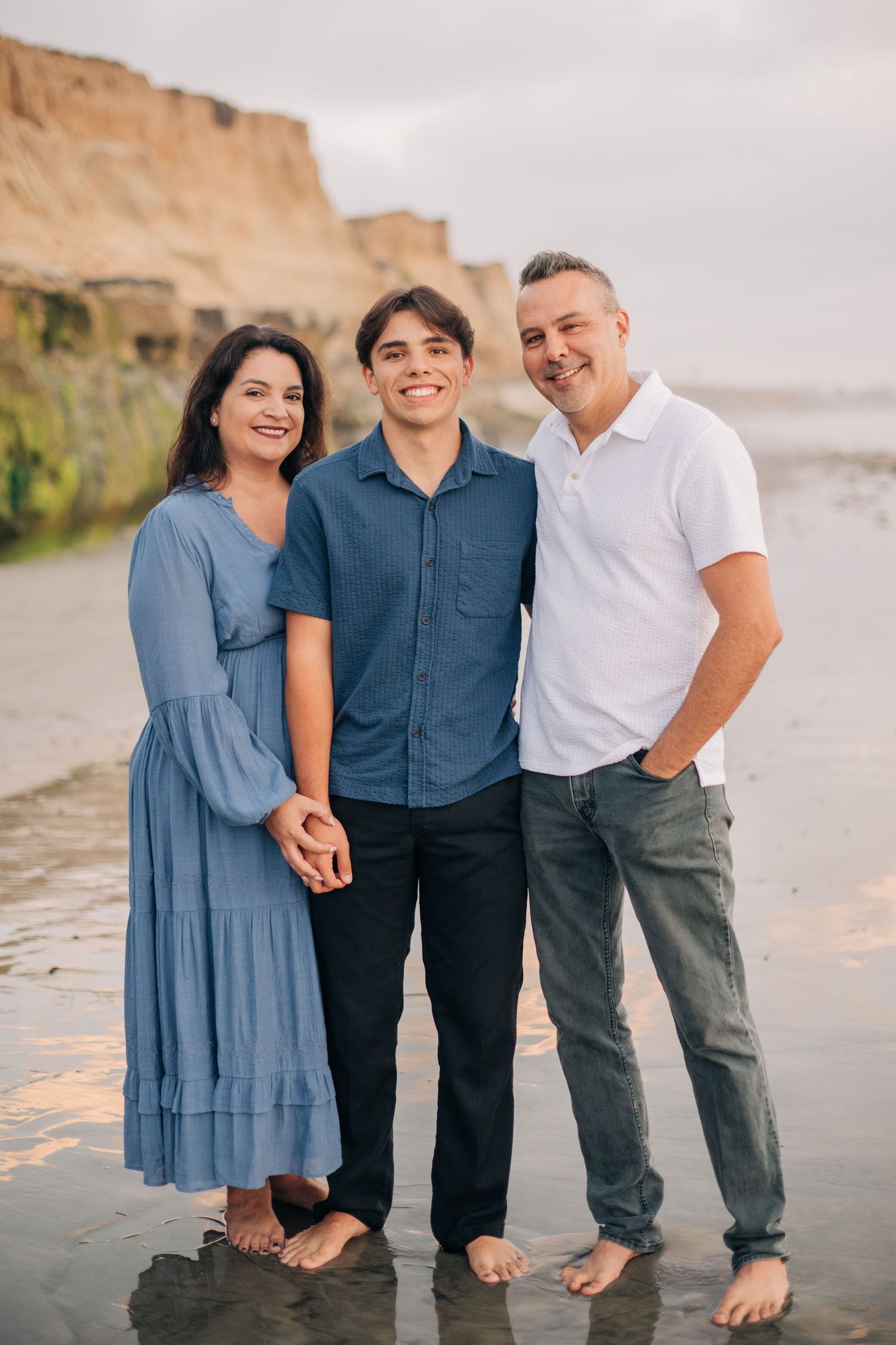 Family of three smiling together on a beach at sunset, with cliffs in the background, wearing blue and white outfits.