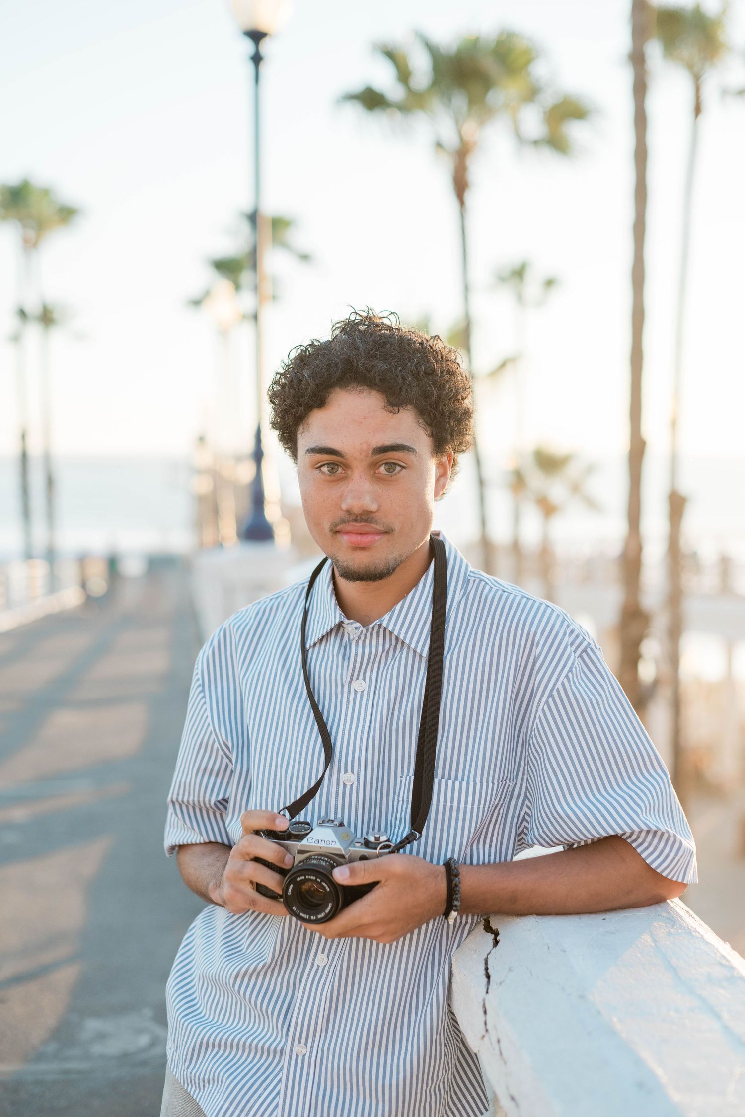 Young man with curly hair holding a vintage film camera on a sunny beachside promenade with palm trees.