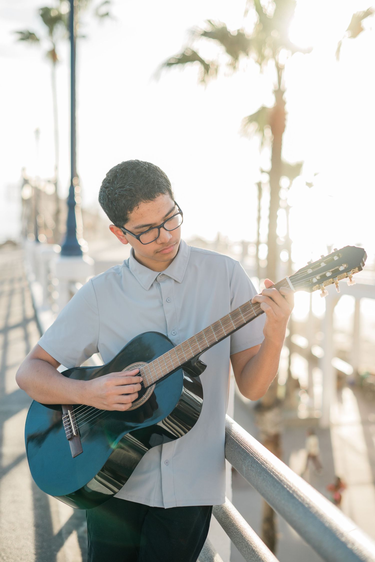 Young man with glasses playing acoustic guitar outdoors near a pier with palm trees in sunny weather.