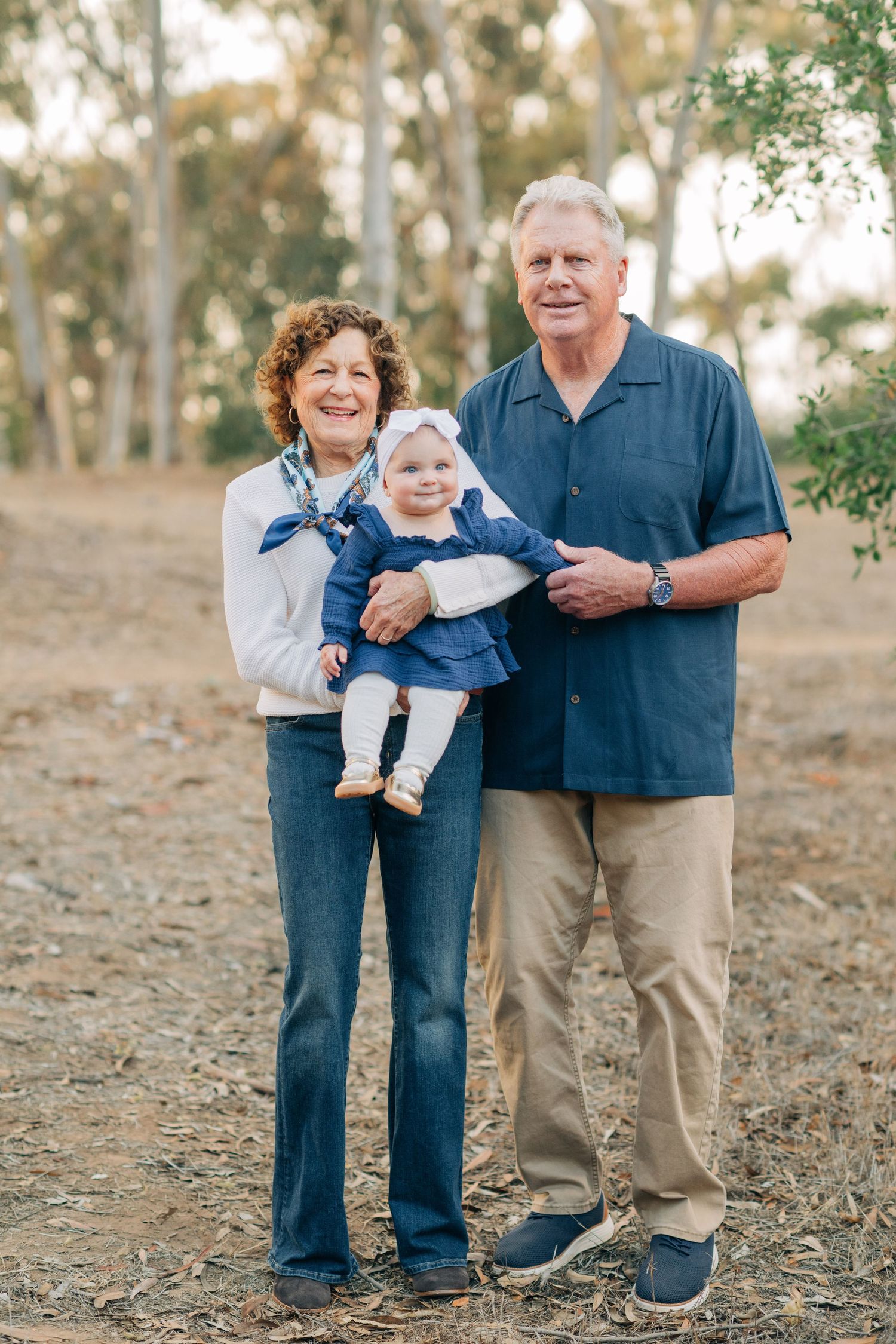 Smiling grandparents holding a happy baby girl outdoors in a park setting with trees in the background.