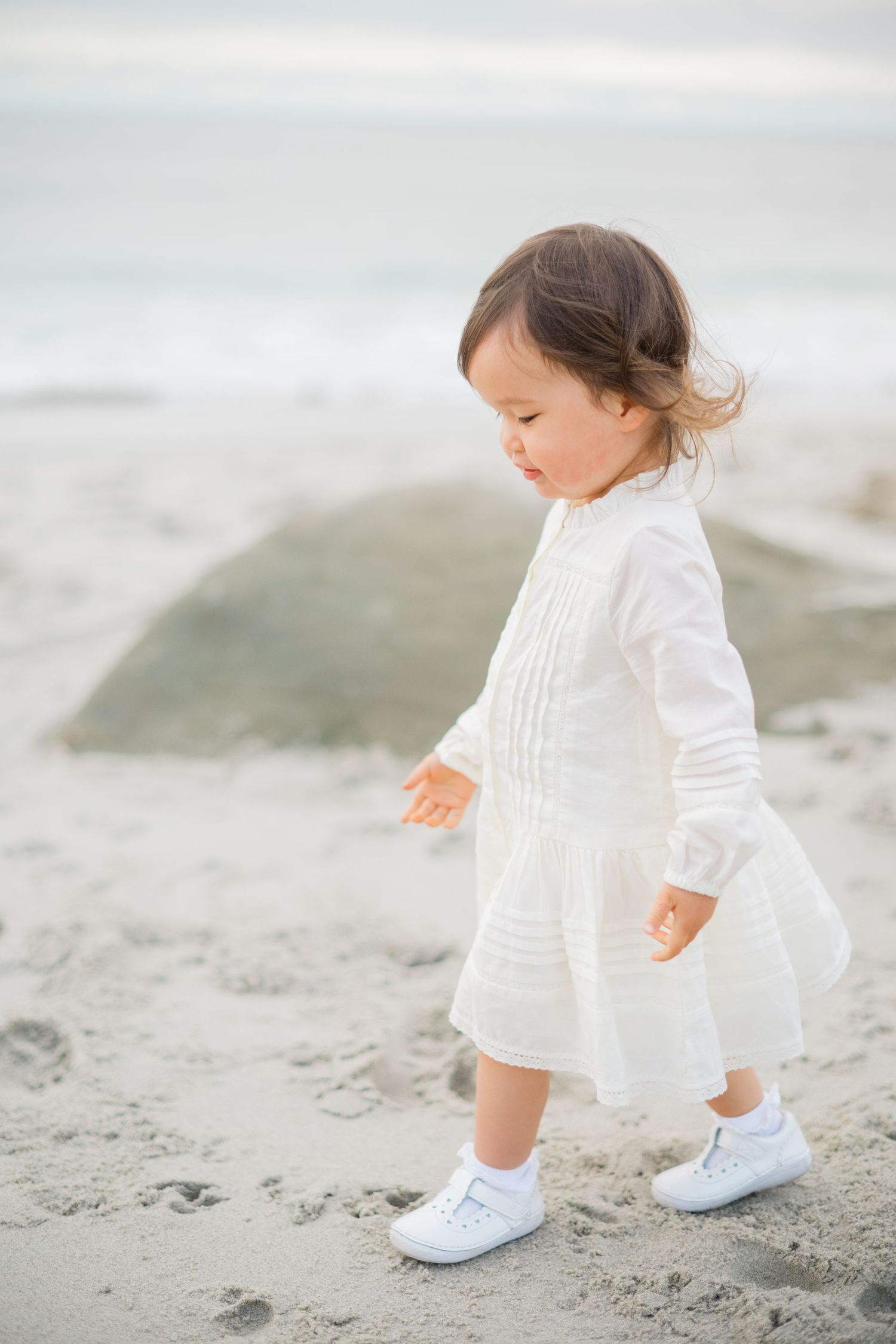 Toddler girl in white dress and shoes walking on sandy beach near ocean waves.
