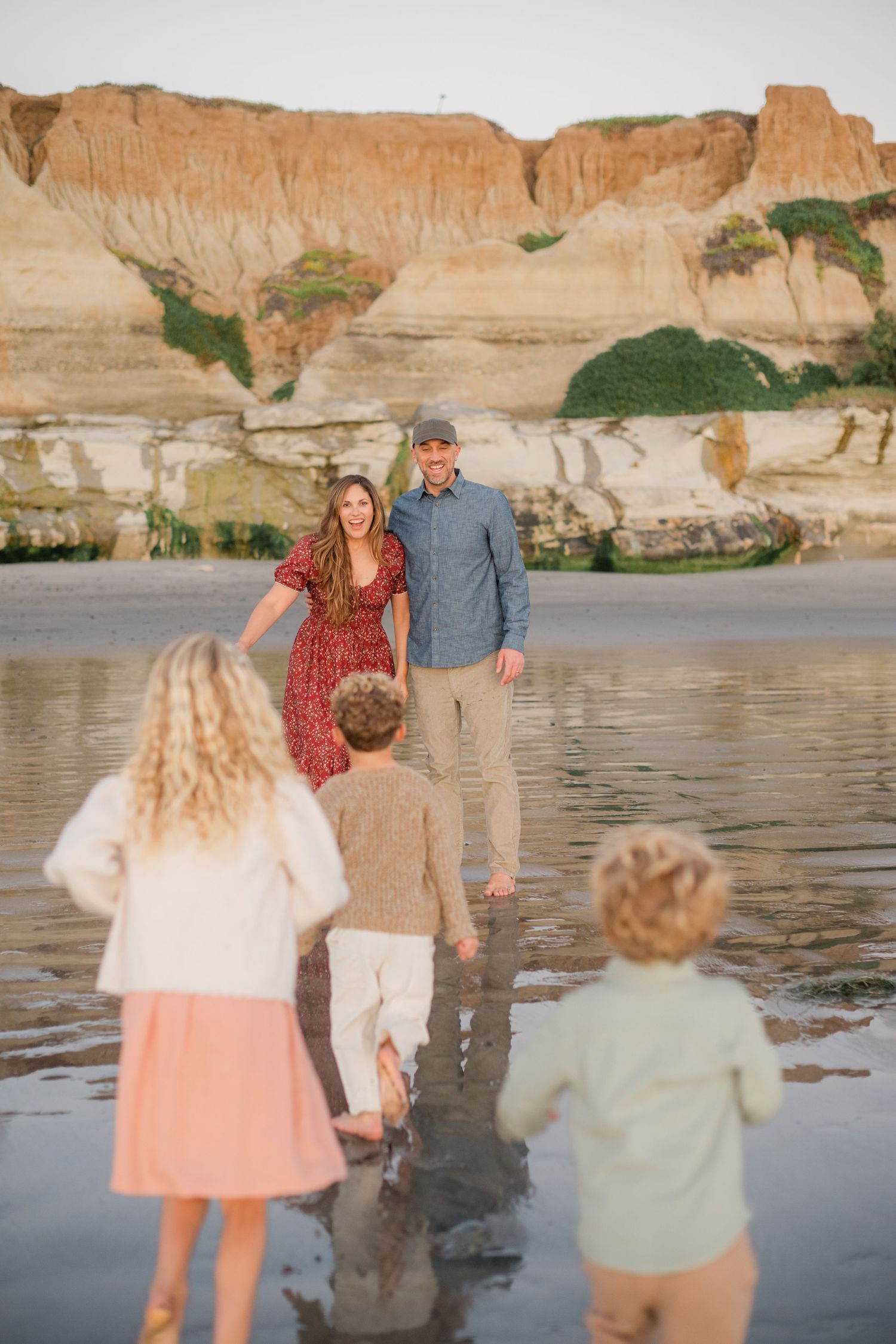 Parents smiling as three children run toward them on a reflective beach with sandstone cliffs.