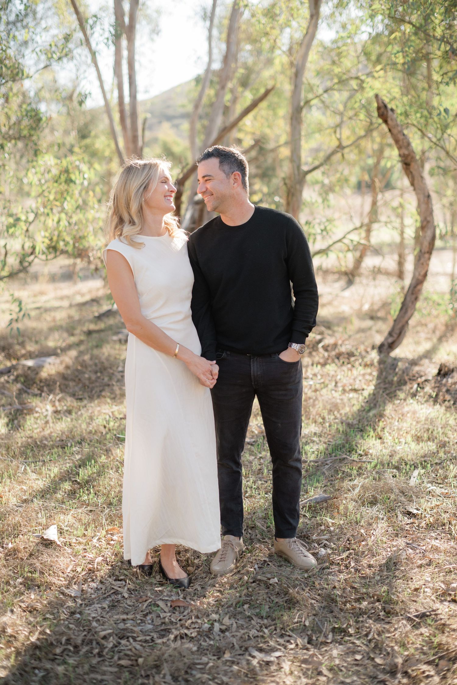 A smiling couple holds hands in a sunlit outdoor setting, woman in white dress, man in black sweater and jeans.