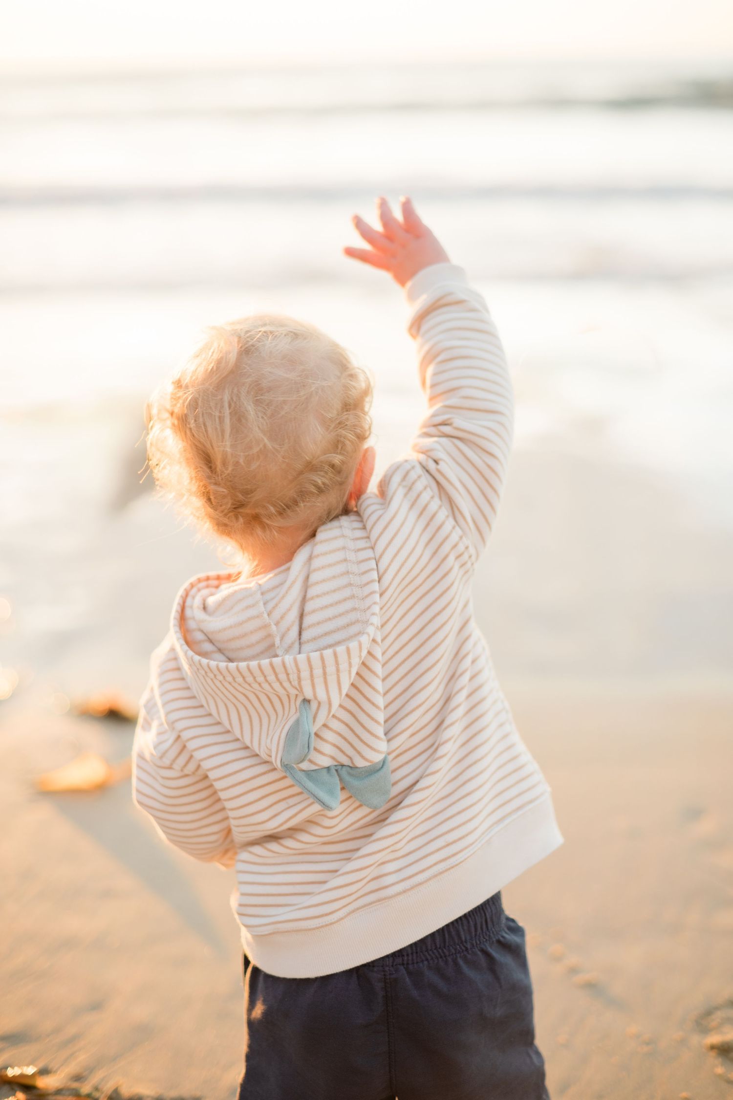 Toddler in striped hoodie reaching toward ocean waves on sunny beach at golden hour.