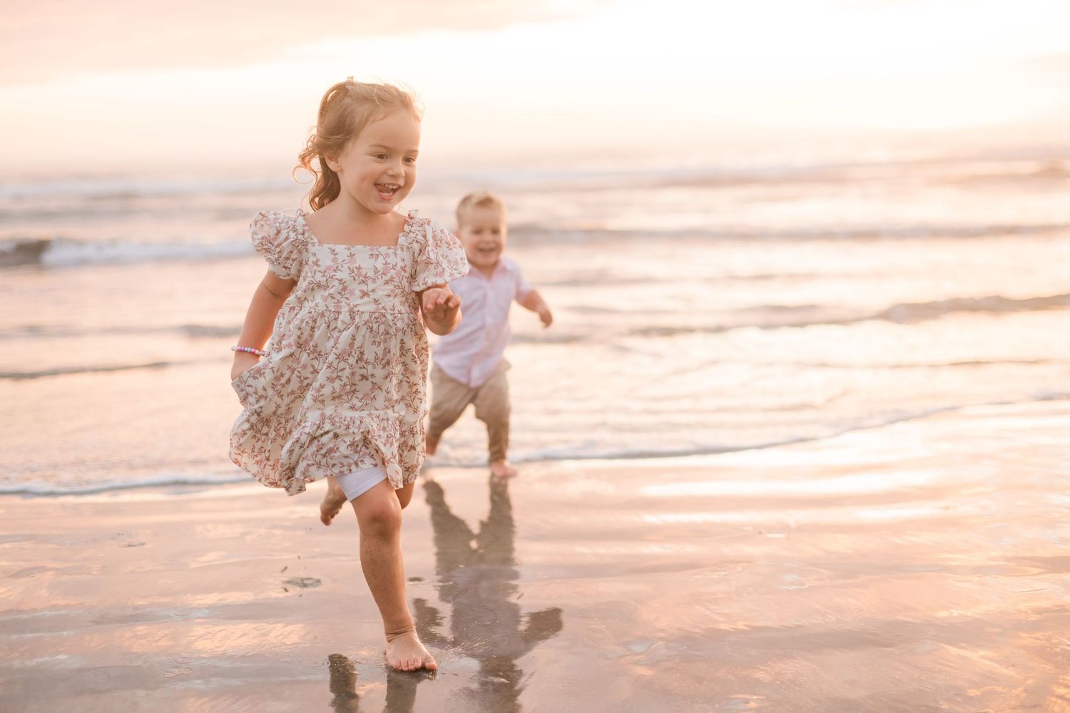 Two young children laughing and running along a sandy beach at sunset, waves gently lapping at their feet.