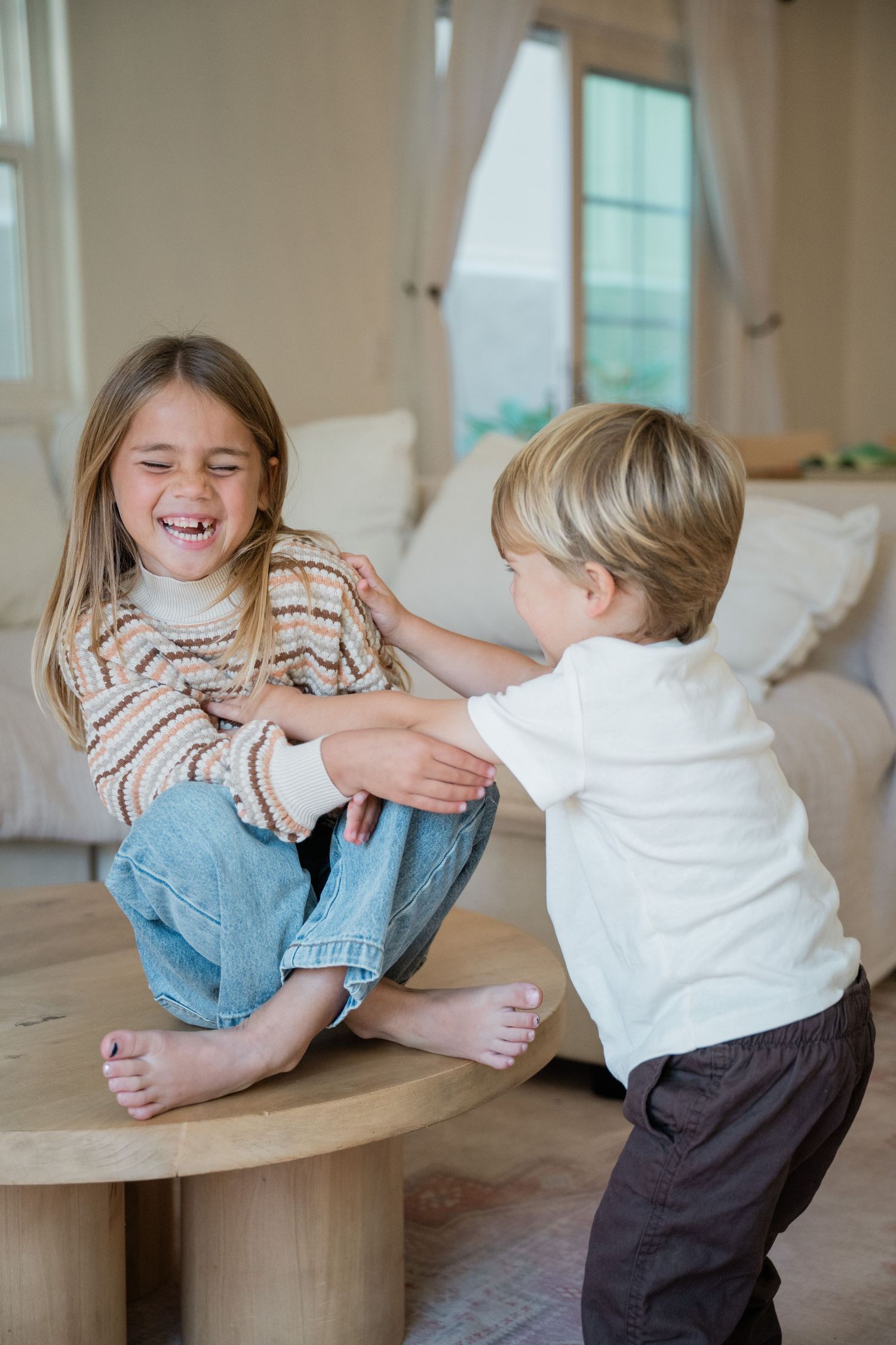 Two barefoot children playing and laughing together on a wooden coffee table in a bright living room.