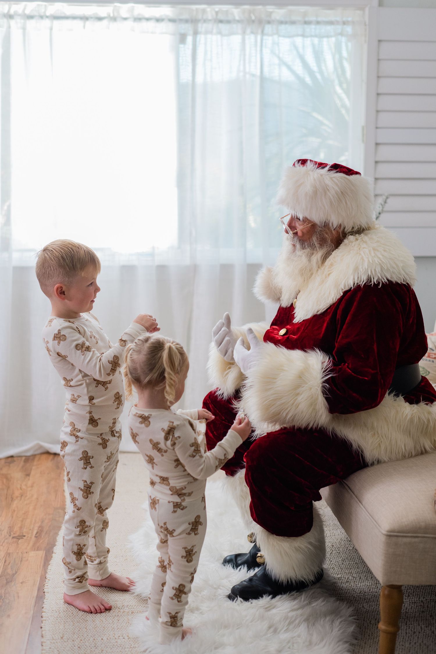 Two children in matching gingerbread pajamas visit Santa Claus seated in a cozy white room during Christmas.