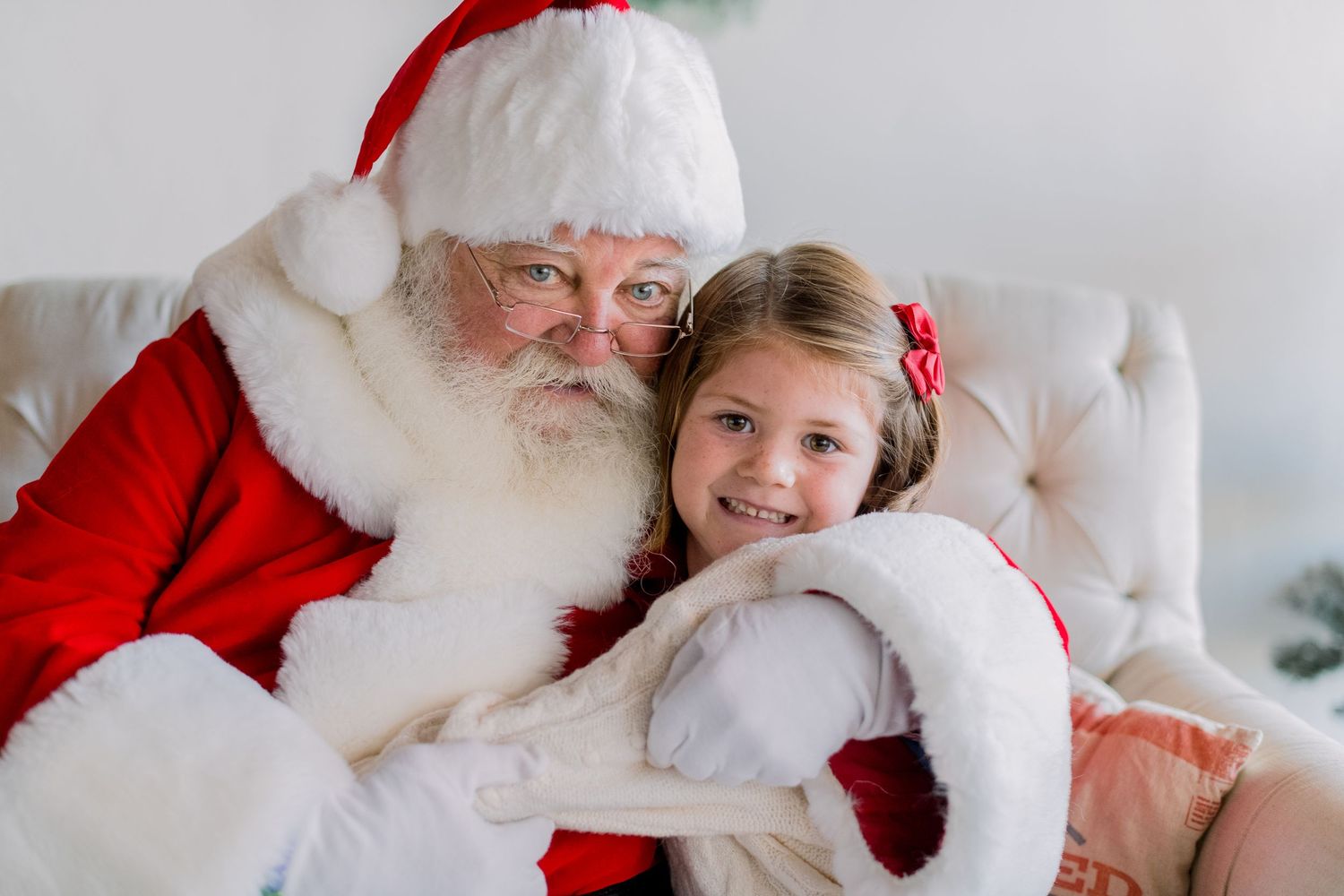A smiling girl in a red bow hugs Santa Claus in a festive holiday portrait photo.