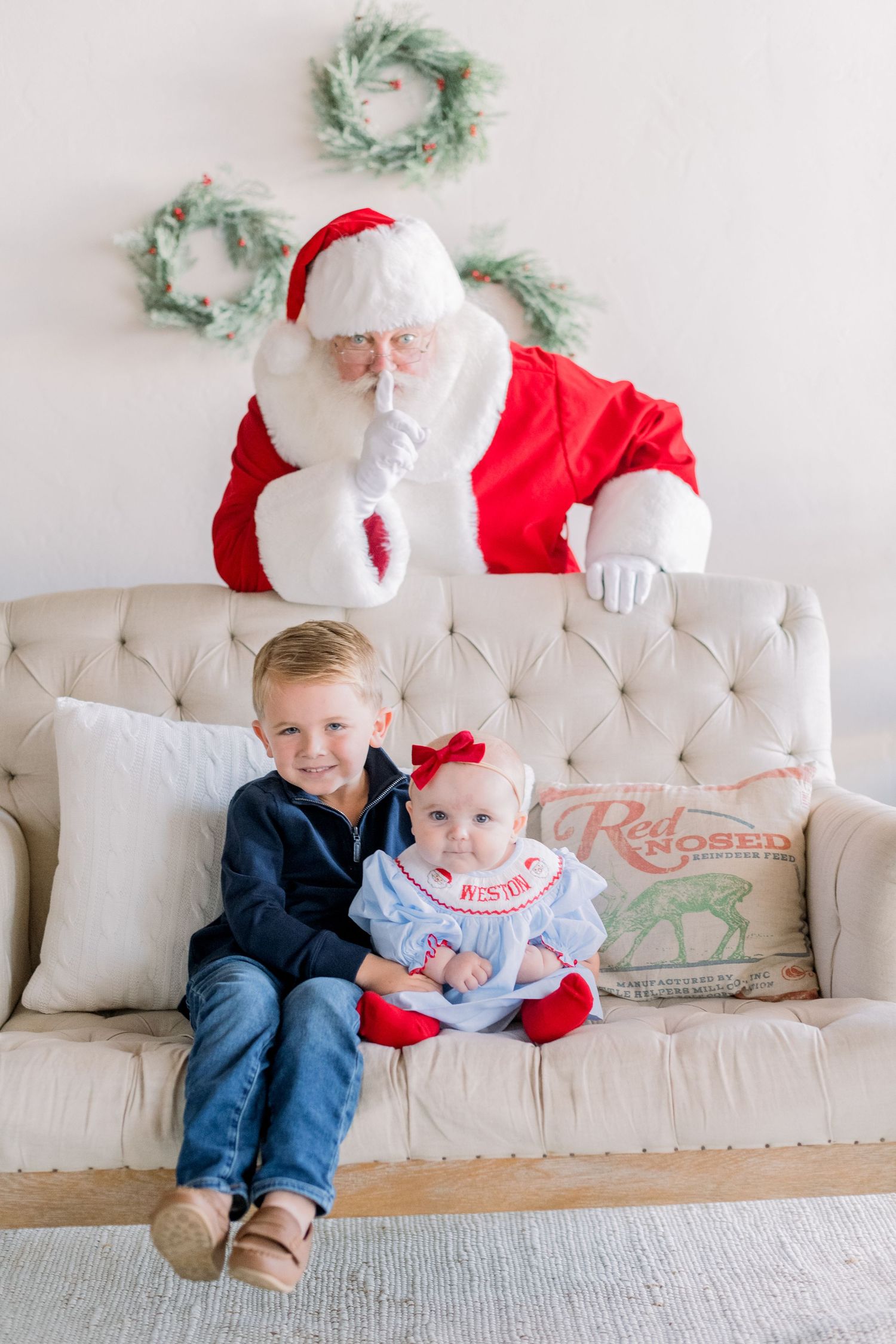Two children sit on a beige tufted sofa with Santa Claus behind them making a shushing gesture, with holiday wreaths on the wall.