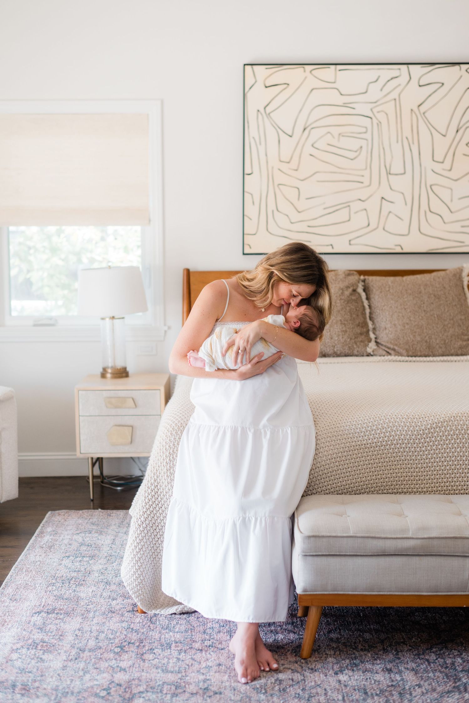 Mother in white dress kissing her newborn baby in bright modern bedroom.